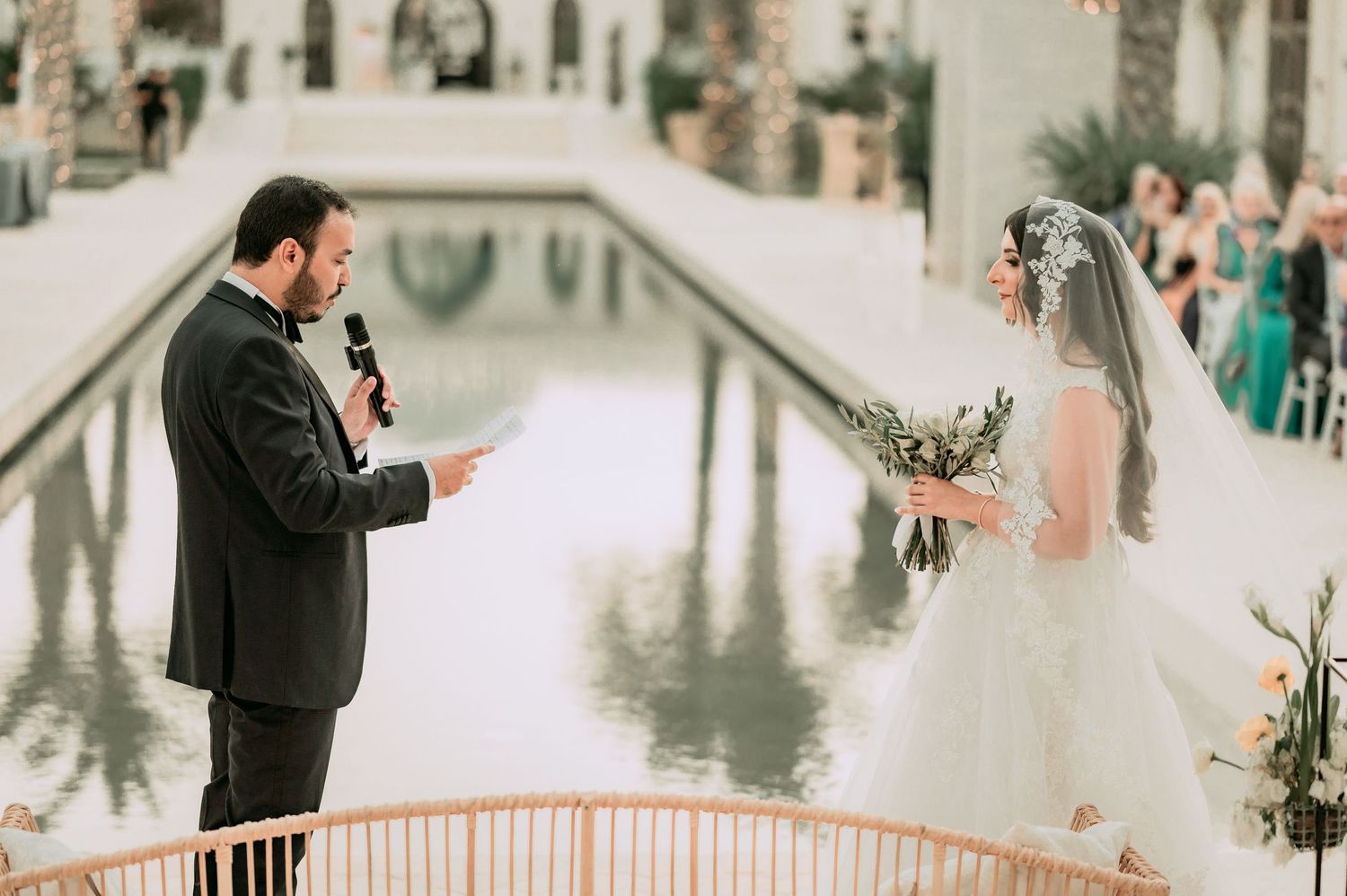Groom reads wedding vows to bride in white ball gown at outdoor ceremony beside a shimmering reflecting pool.