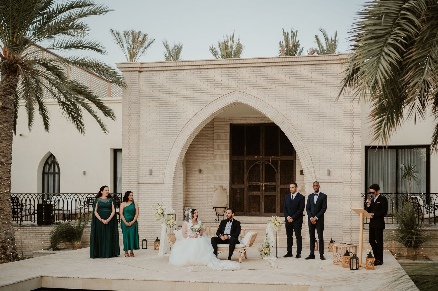 Wedding ceremony with bride, groom, bridesmaids in green, and groomsmen before an arched stone building.