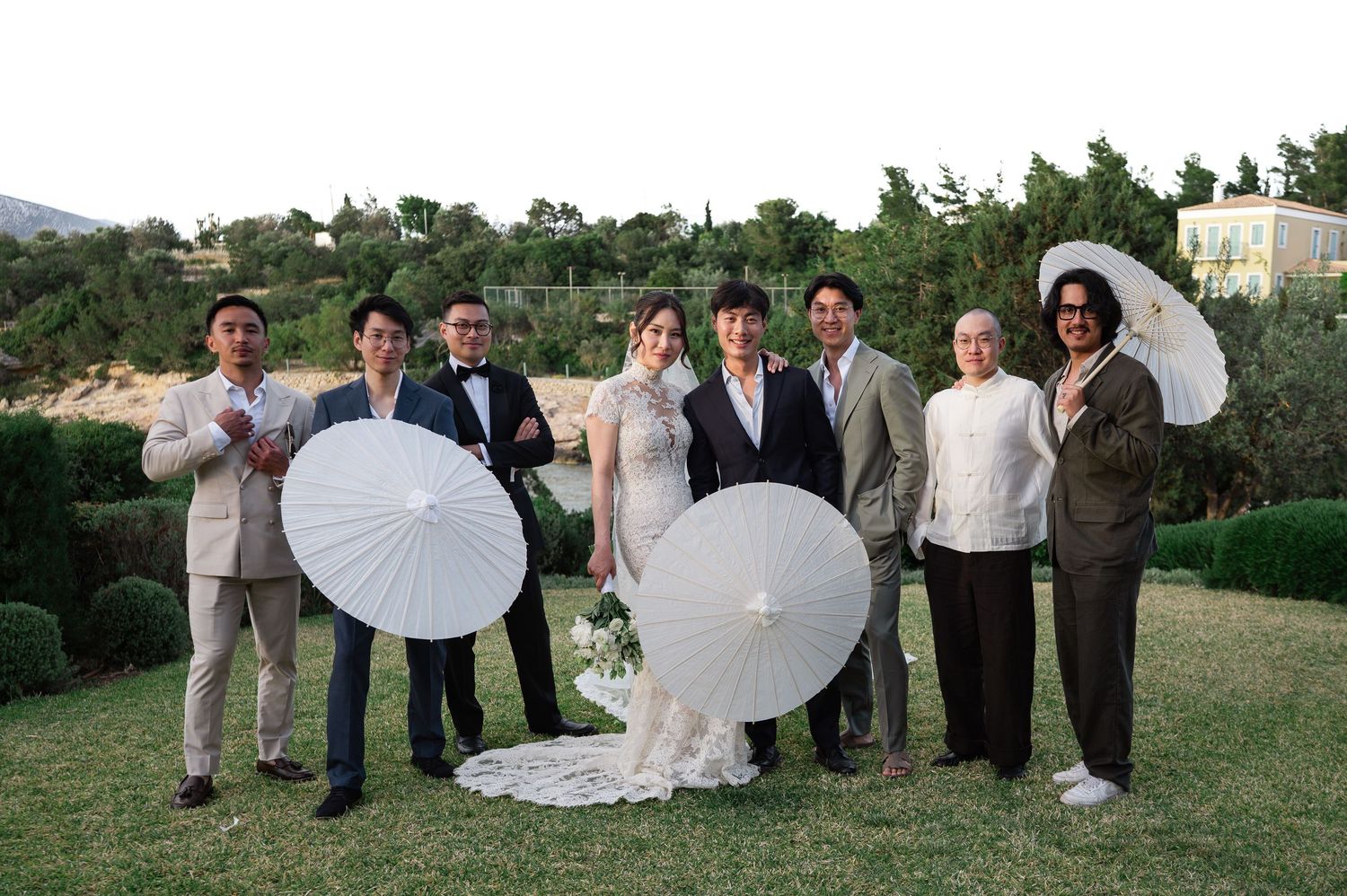 Wedding party poses with white parasols on lawn with groom, bride, and guests in formal attire.