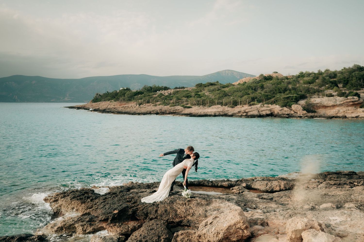 Groom dips bride dramatically on rocky seaside cliffs with stunning turquoise water and mountains behind.