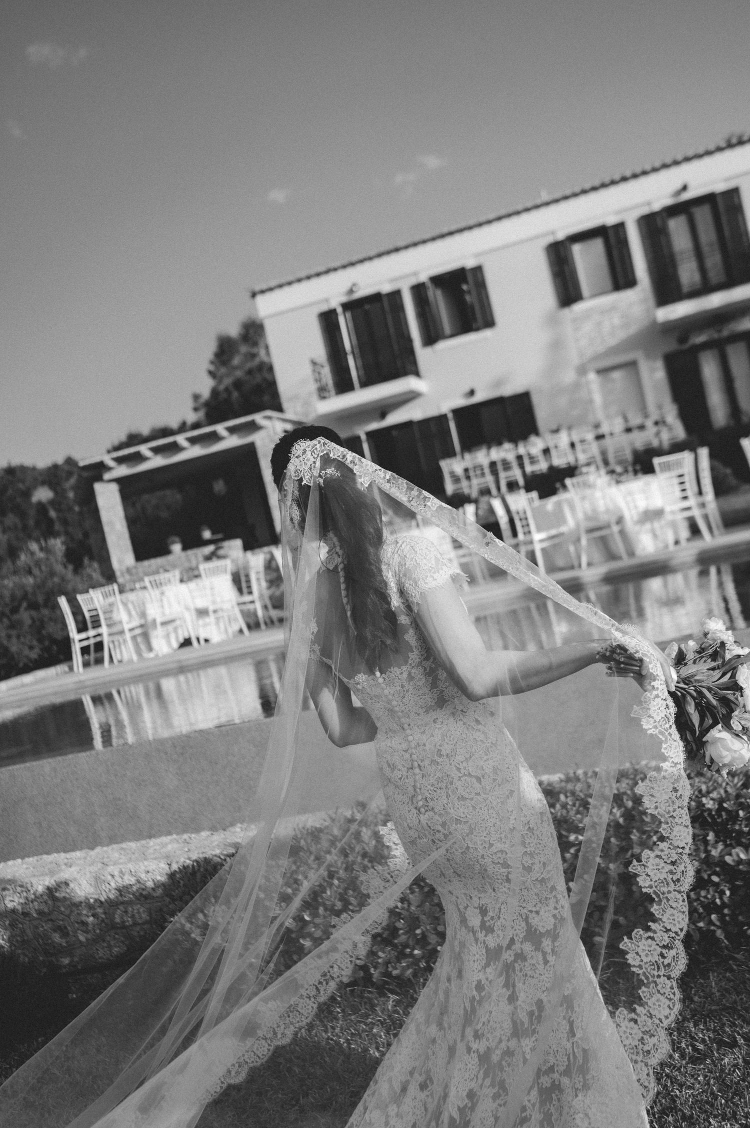 Bride in lace wedding dress with flowing veil caught in the wind at an outdoor venue.