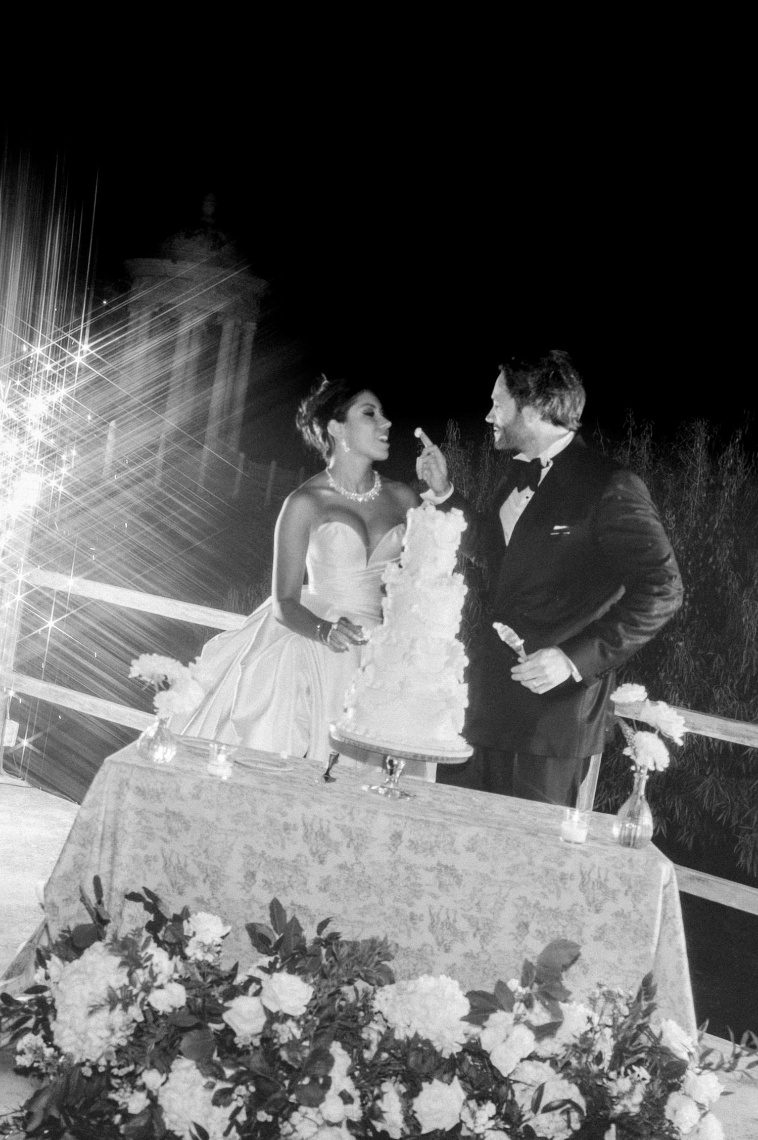 Bride and groom share a tender cake-cutting moment surrounded by flowers and sparkling lights at night.