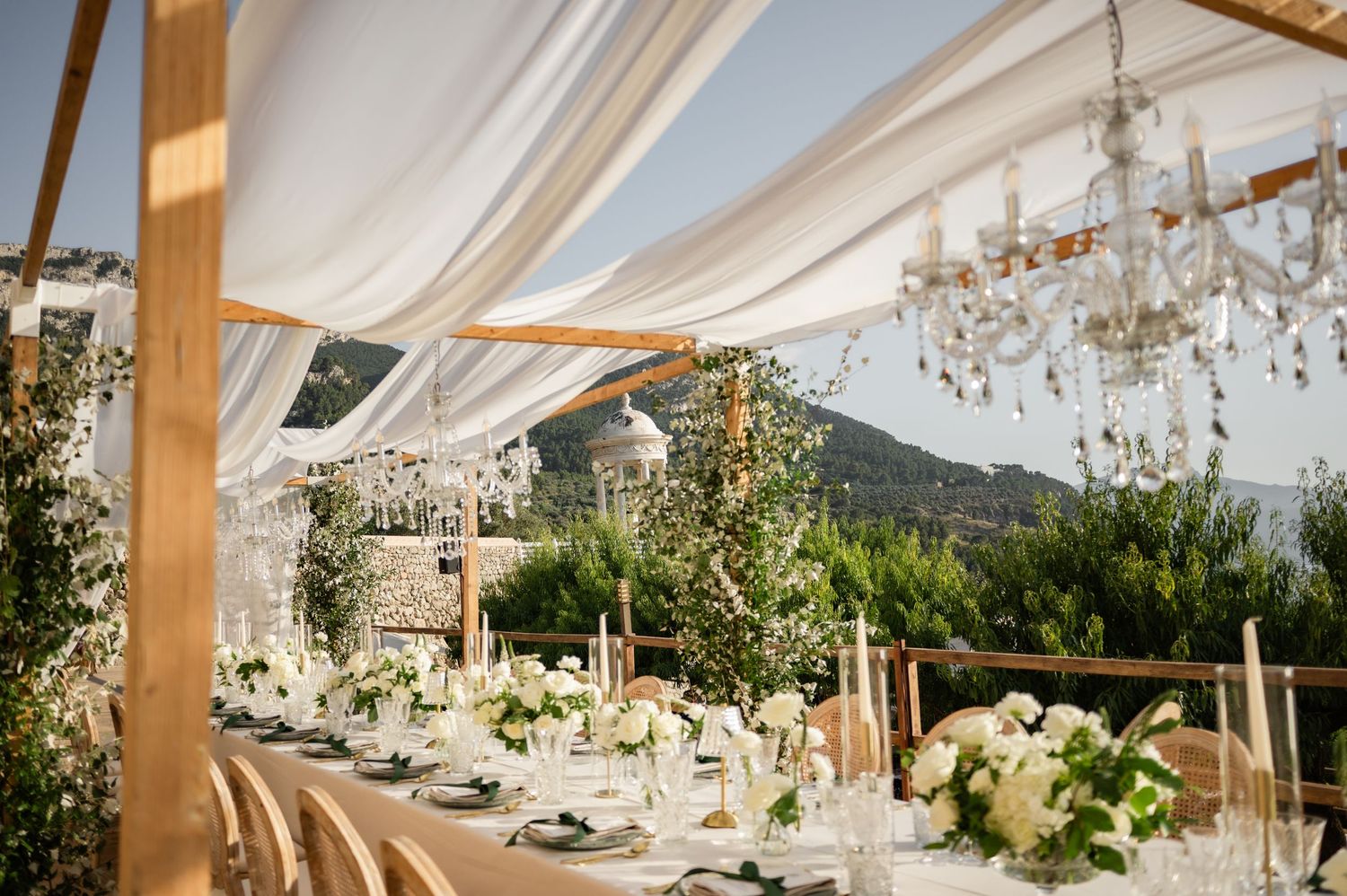 Luxurious outdoor wedding reception under white draped canopy with crystal chandeliers, white florals, and mountain views.