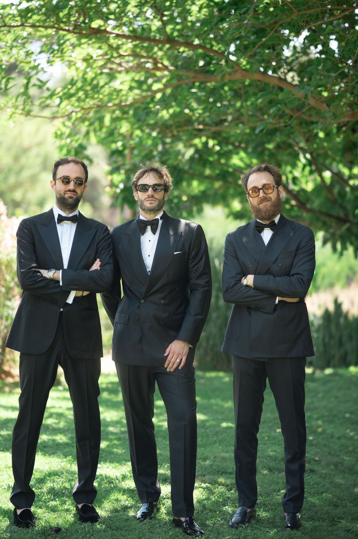 Three groomsmen in black tuxedos and sunglasses pose with crossed arms outdoors under green trees.