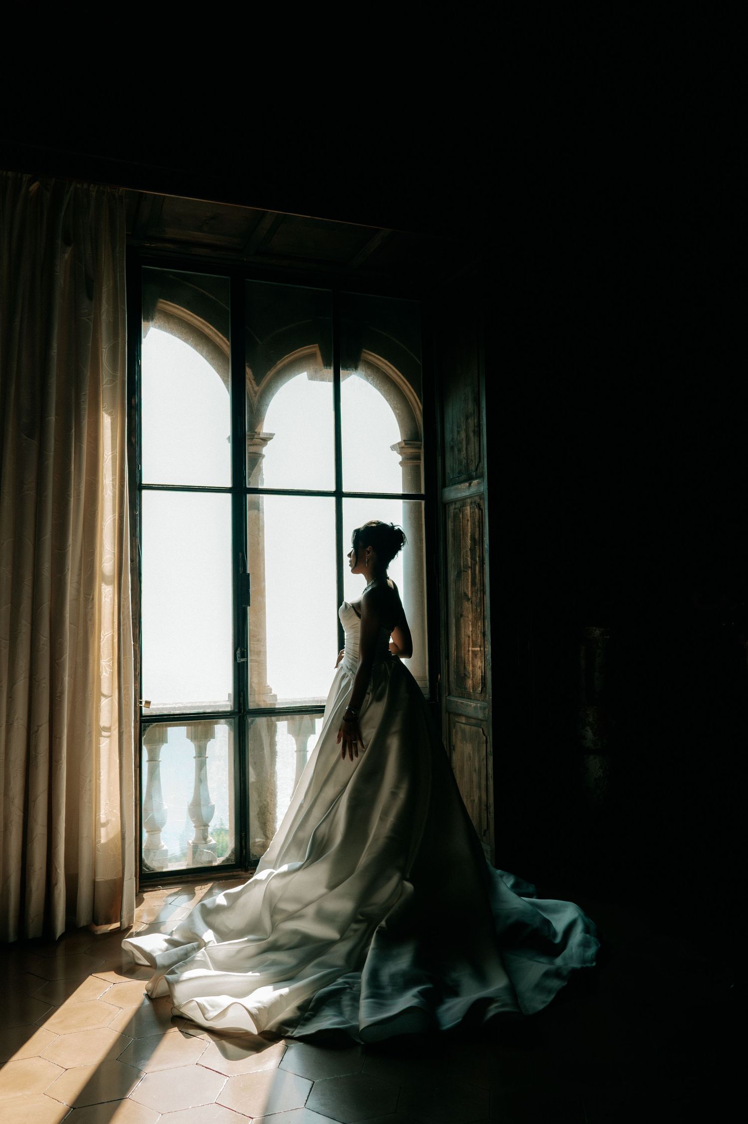 Silhouette of bride in dramatic white gown with long train standing before tall arched window.