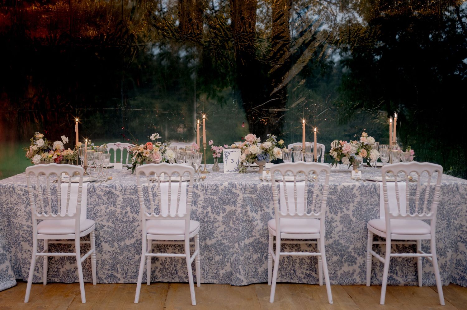 Elegant wedding head table with blue toile tablecloth, white Chiavari chairs, taper candles, and floral arrangements.