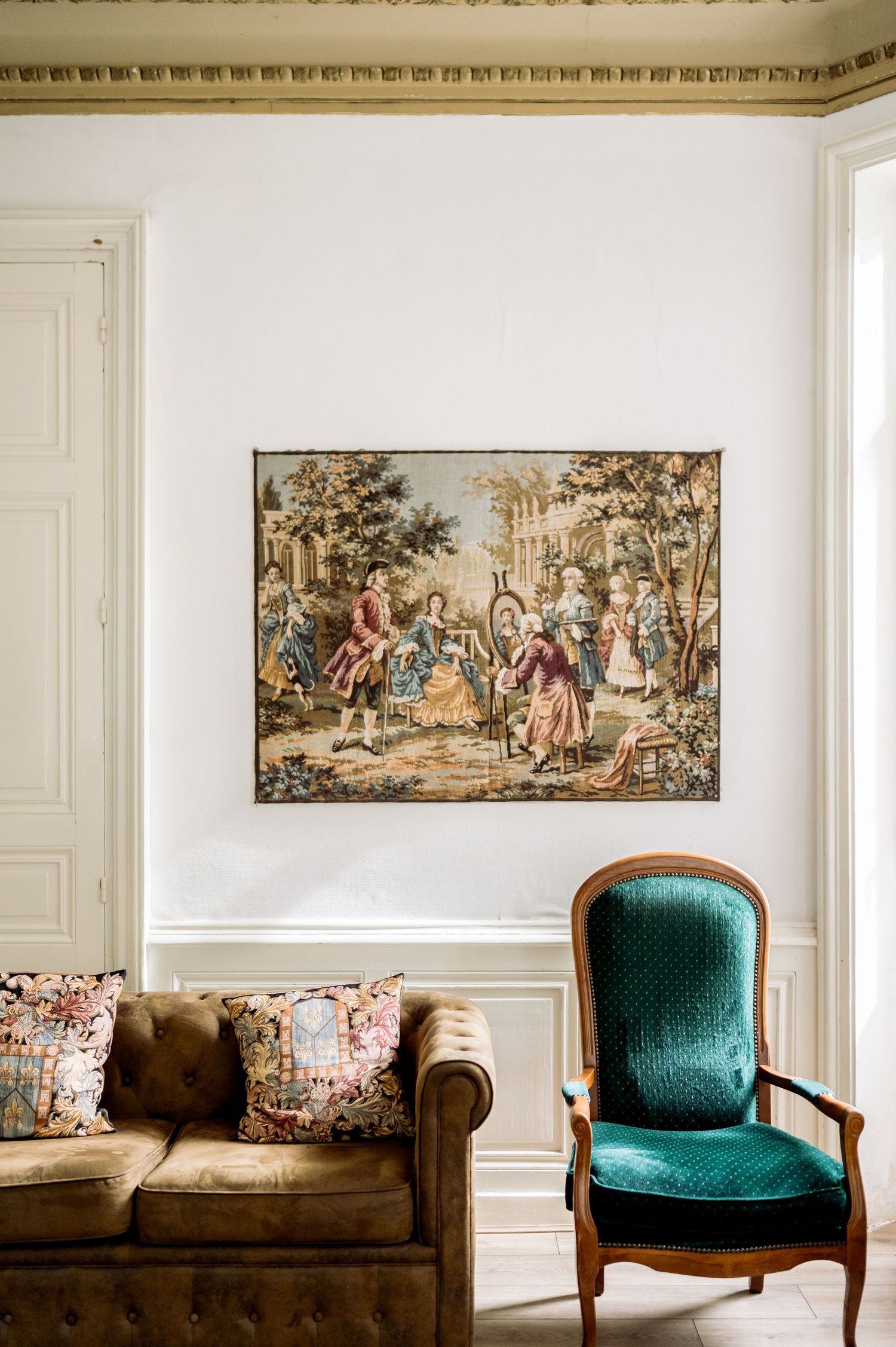 Classic French interior with Aubusson tapestry, teal velvet armchair, and vintage leather sofa with floral cushions.