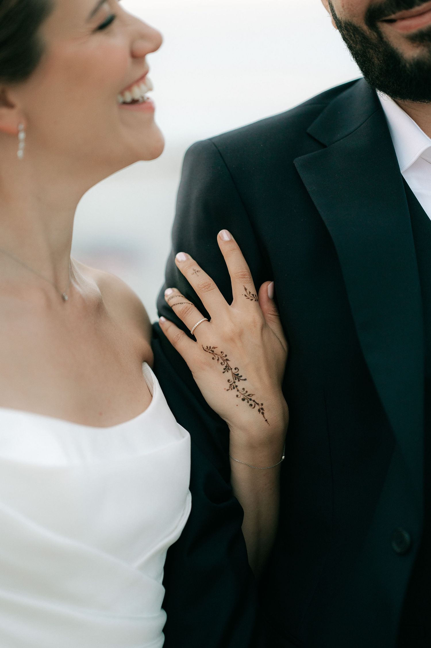 Smiling bride with henna hand art holds grooms arm in dark suit at wedding.