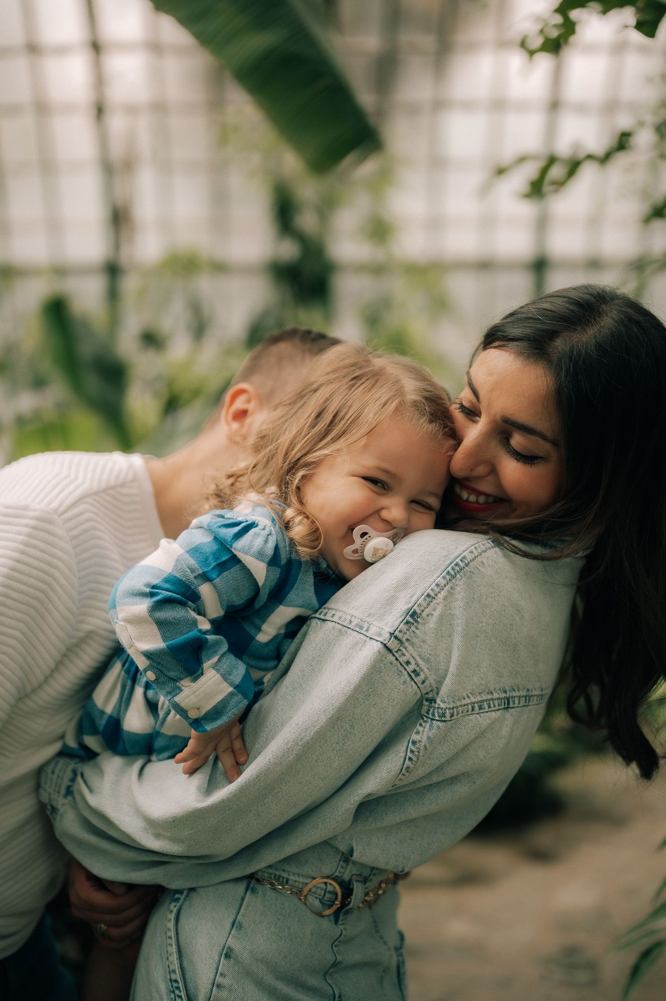 Happy family moment as parents embrace their smiling toddler with a pacifier in a greenhouse.