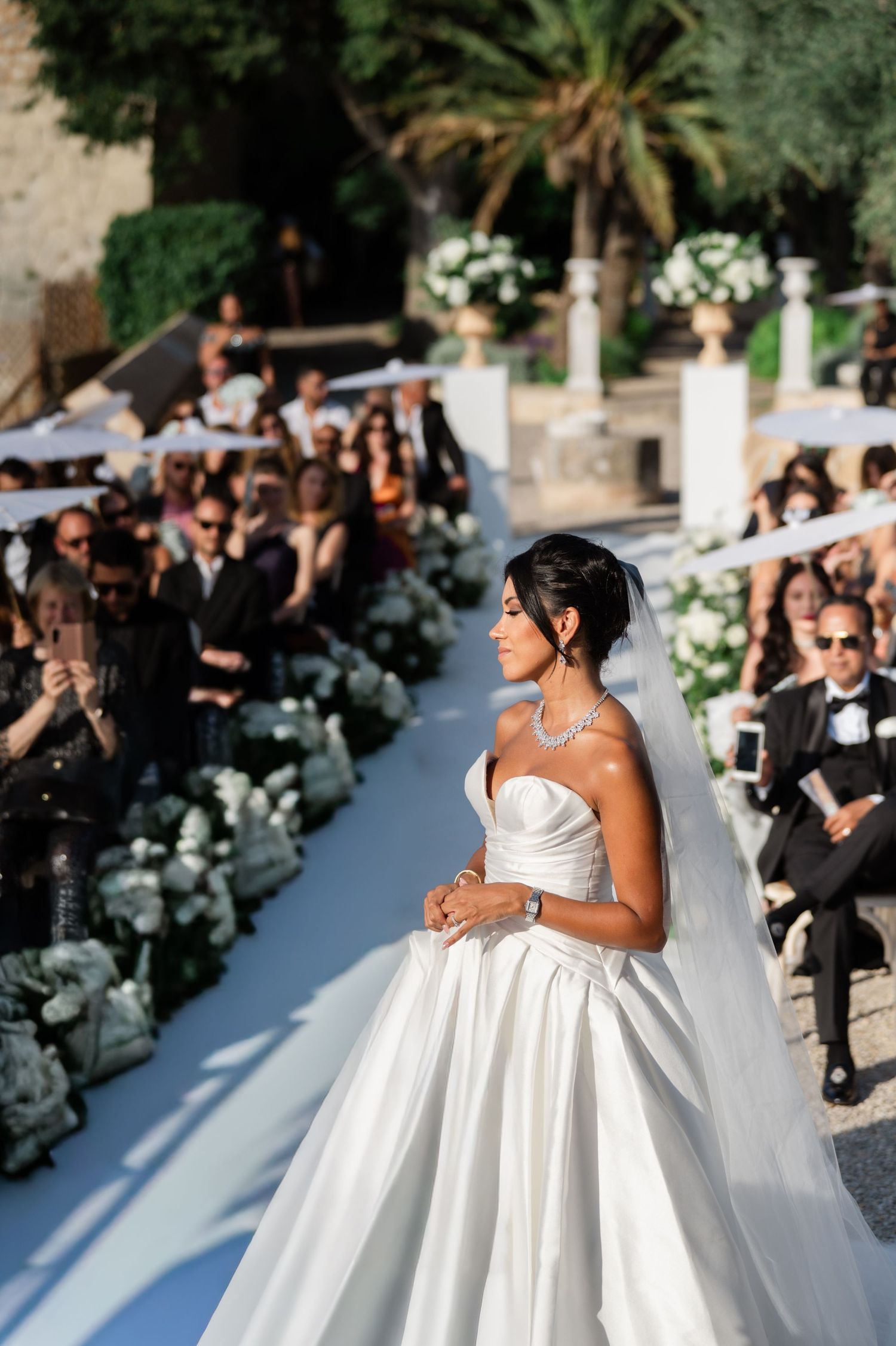 Elegant bride in a white ball gown and veil walks down the aisle at a luxurious outdoor wedding.