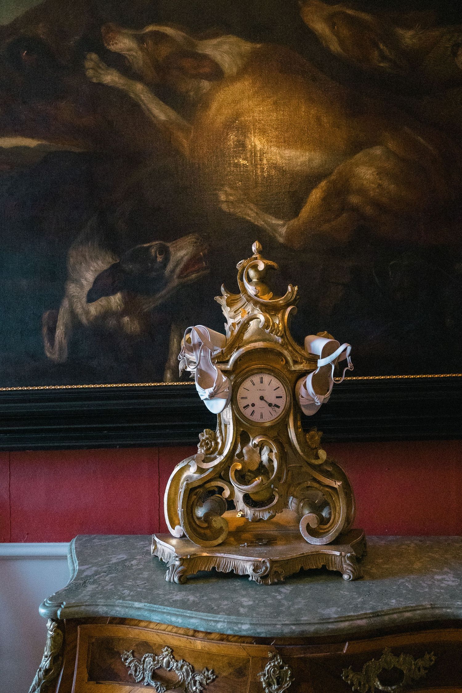 Ornate gold baroque mantel clock with white shoes on marble dresser before dark oil painting.