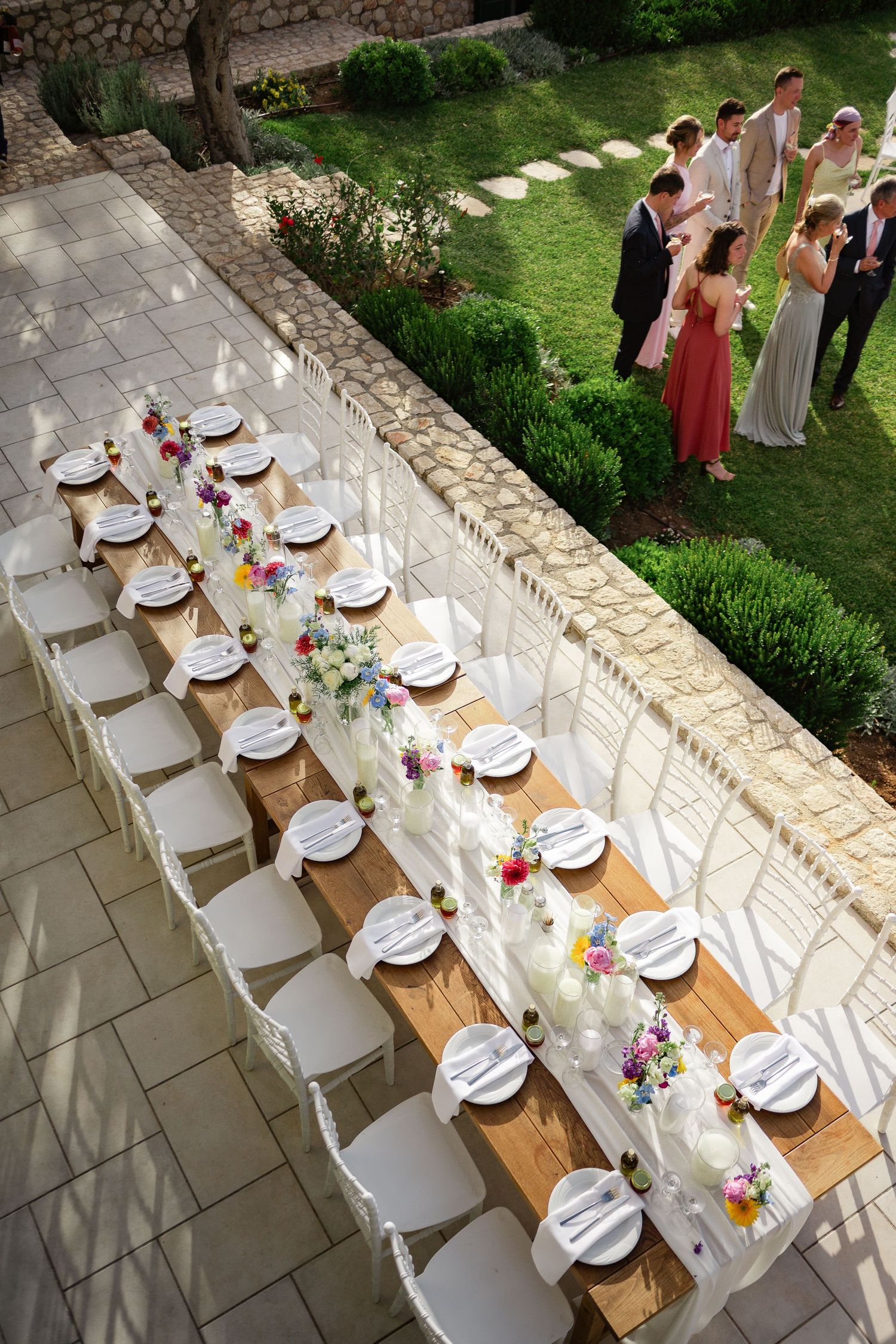 Aerial view of elegant outdoor wedding reception with long wooden table, white chairs, and colorful floral centerpieces.