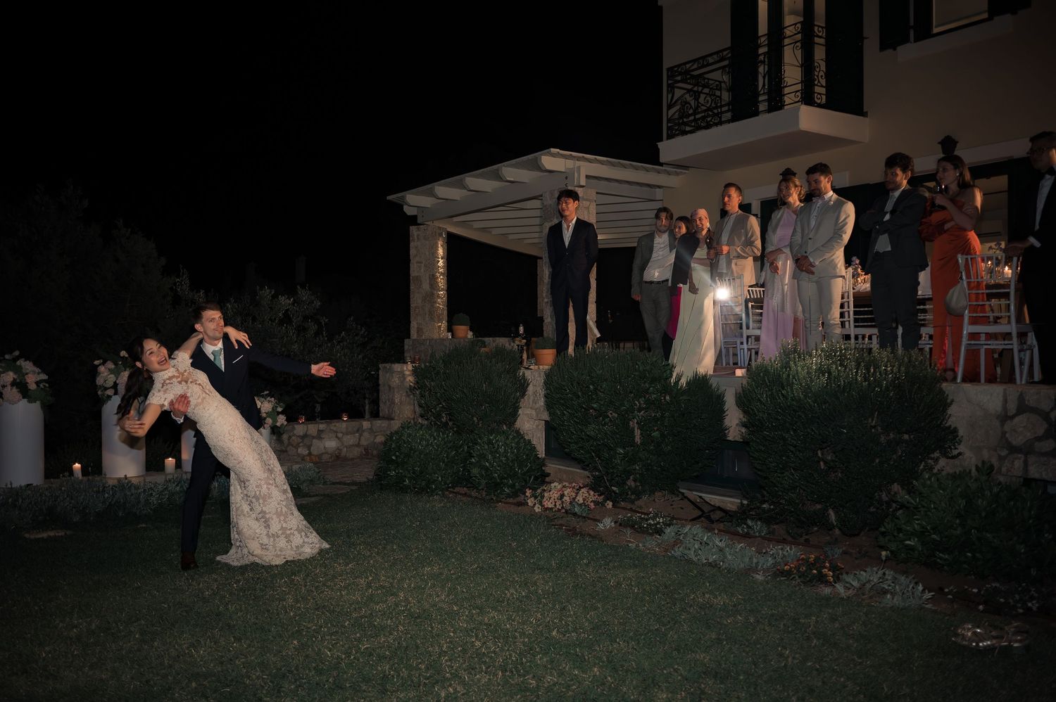 Bride and groom dipping during first dance while wedding guests watch from venue steps at night.