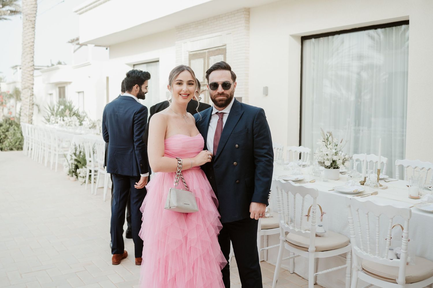 Couple posing at wedding reception, woman in pink ruffled gown and man in navy suit.