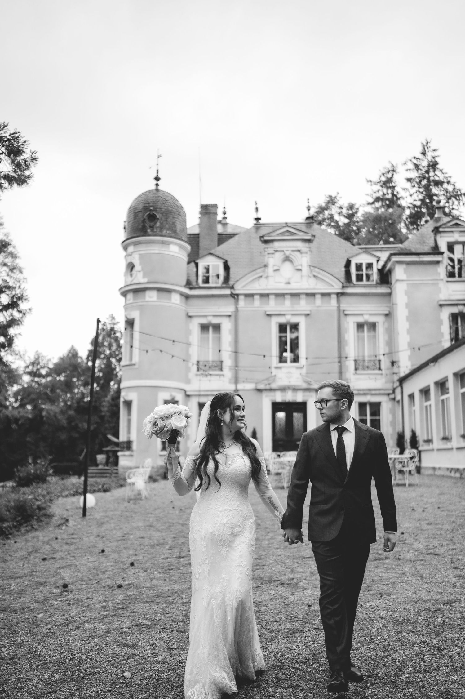 Black and white photo of bride and groom walking hand in hand before a French chateau.