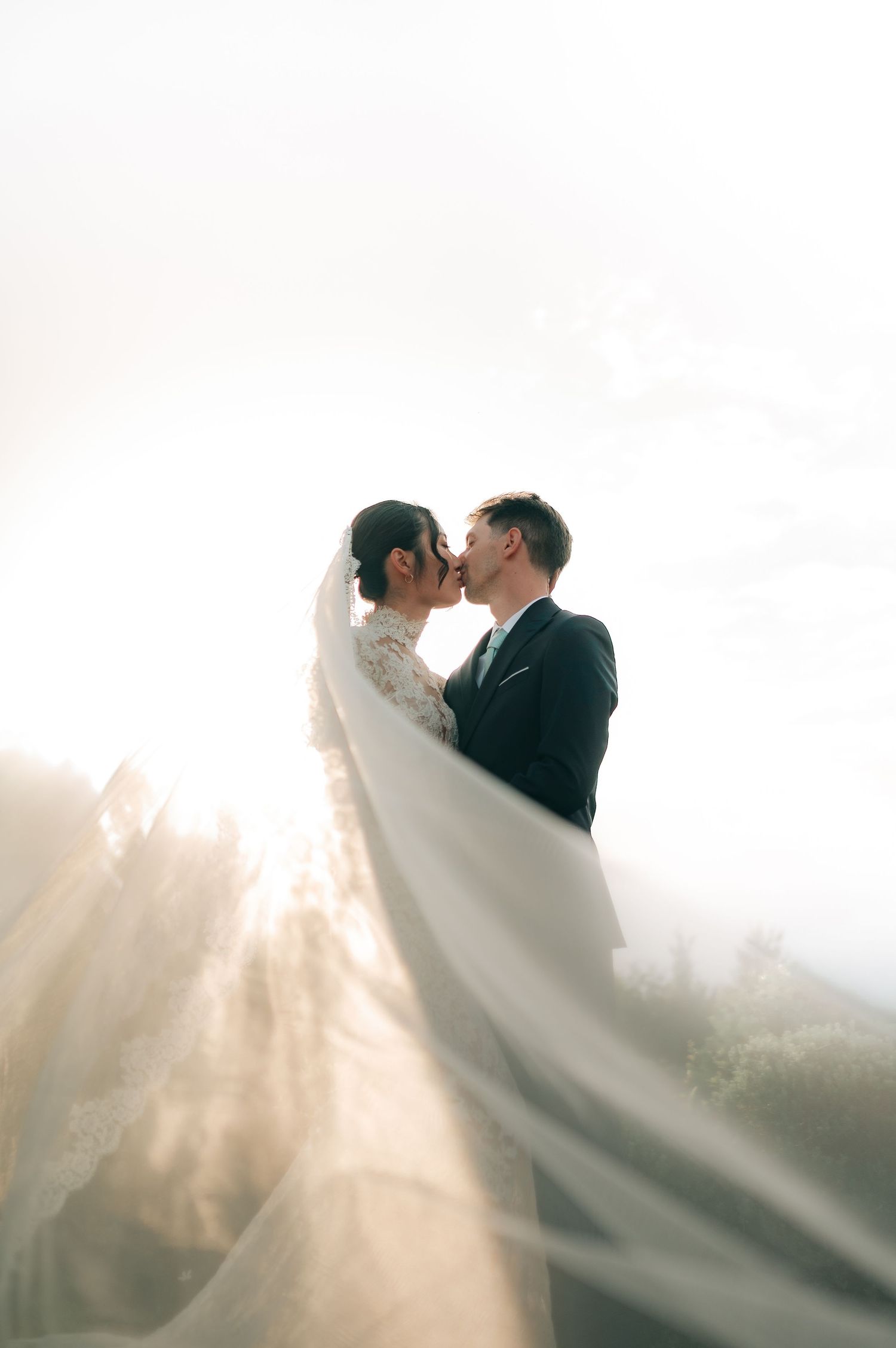 Bride and groom share a kiss backlit by sunlight with veil flowing dramatically in breeze.