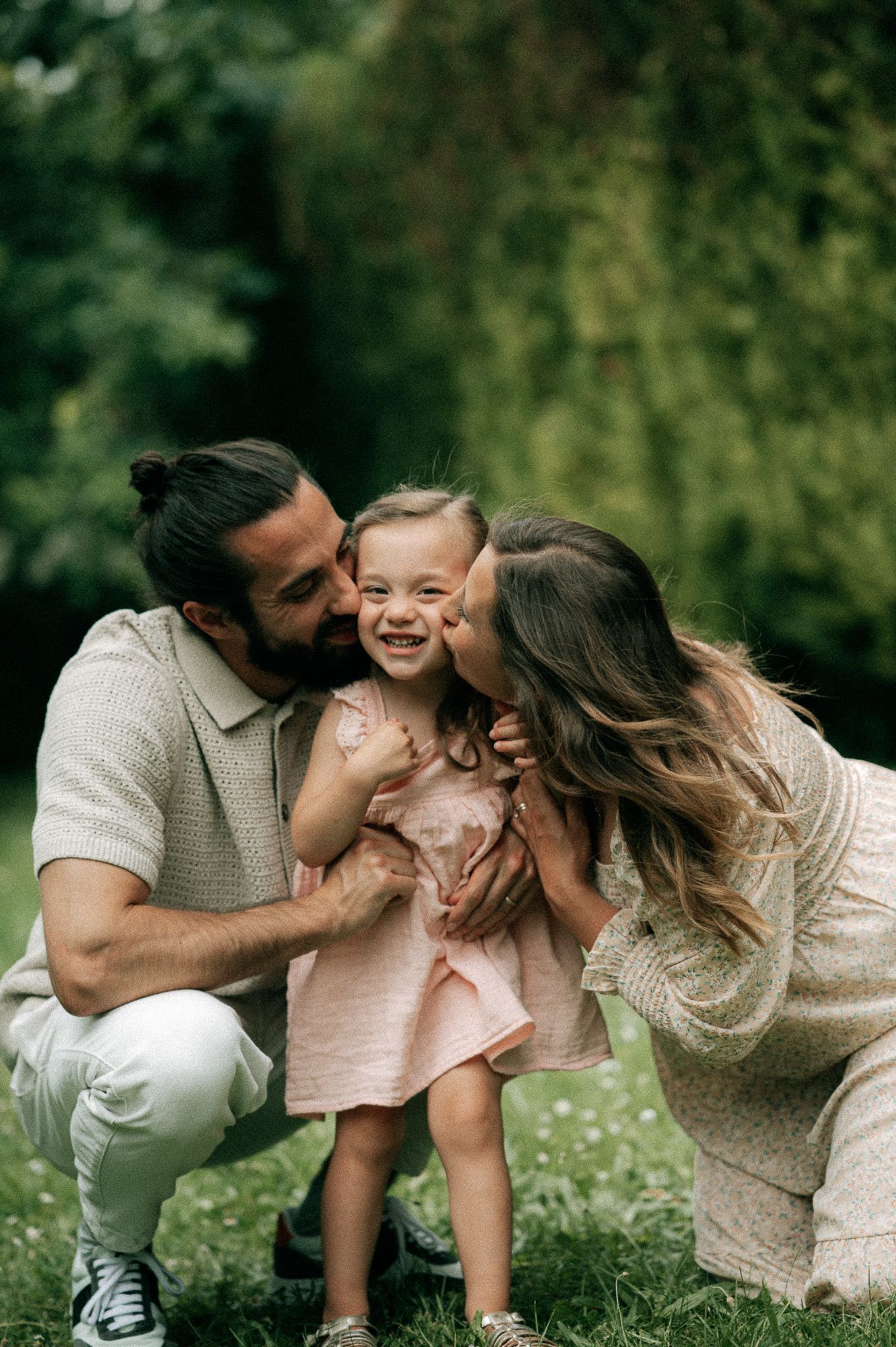 Parents kissing their laughing toddler girl on cheeks outdoors in green park setting.