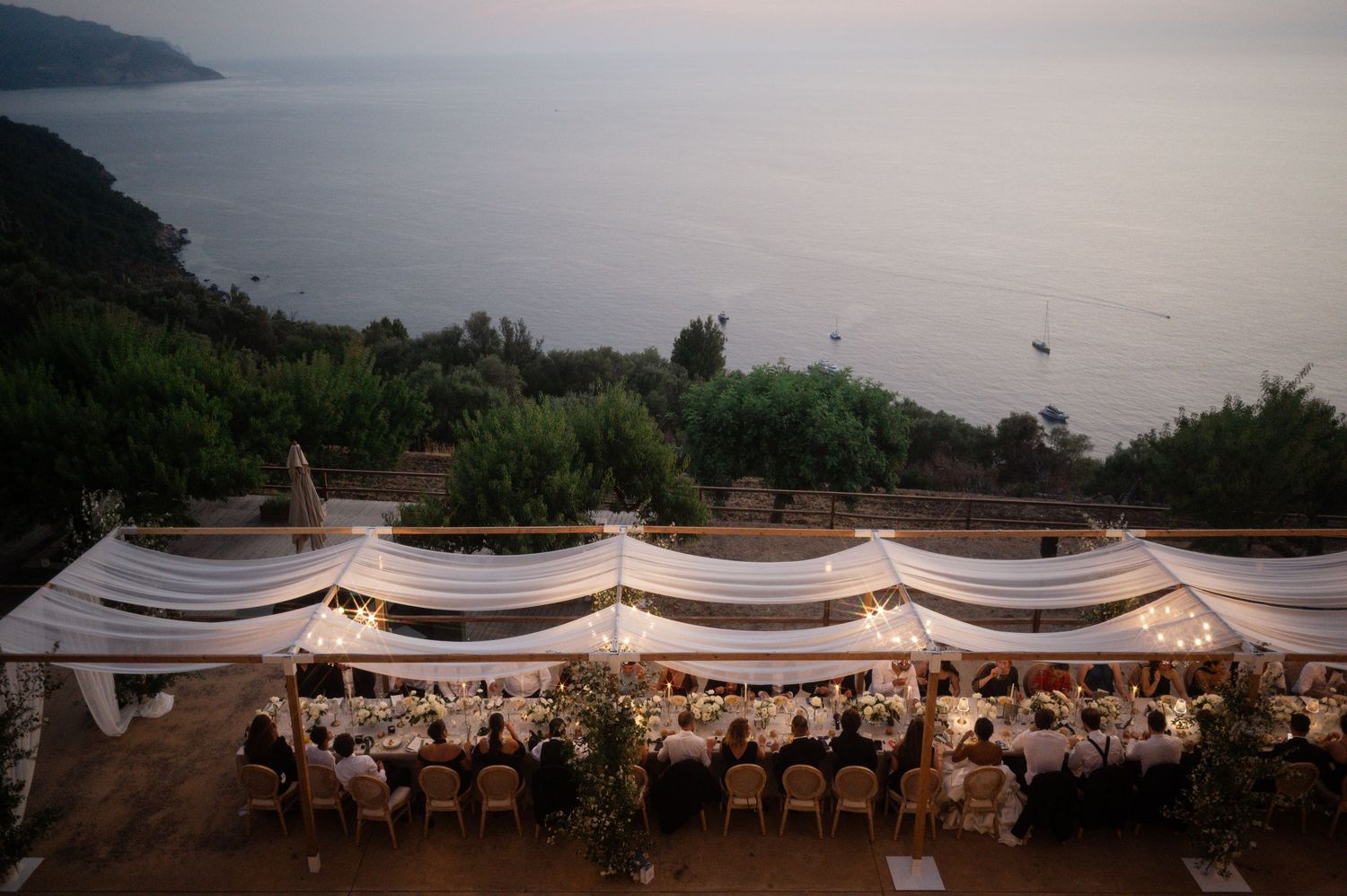Aerial view of an elegant outdoor wedding reception with white draped tables overlooking a stunning coastal landscape.