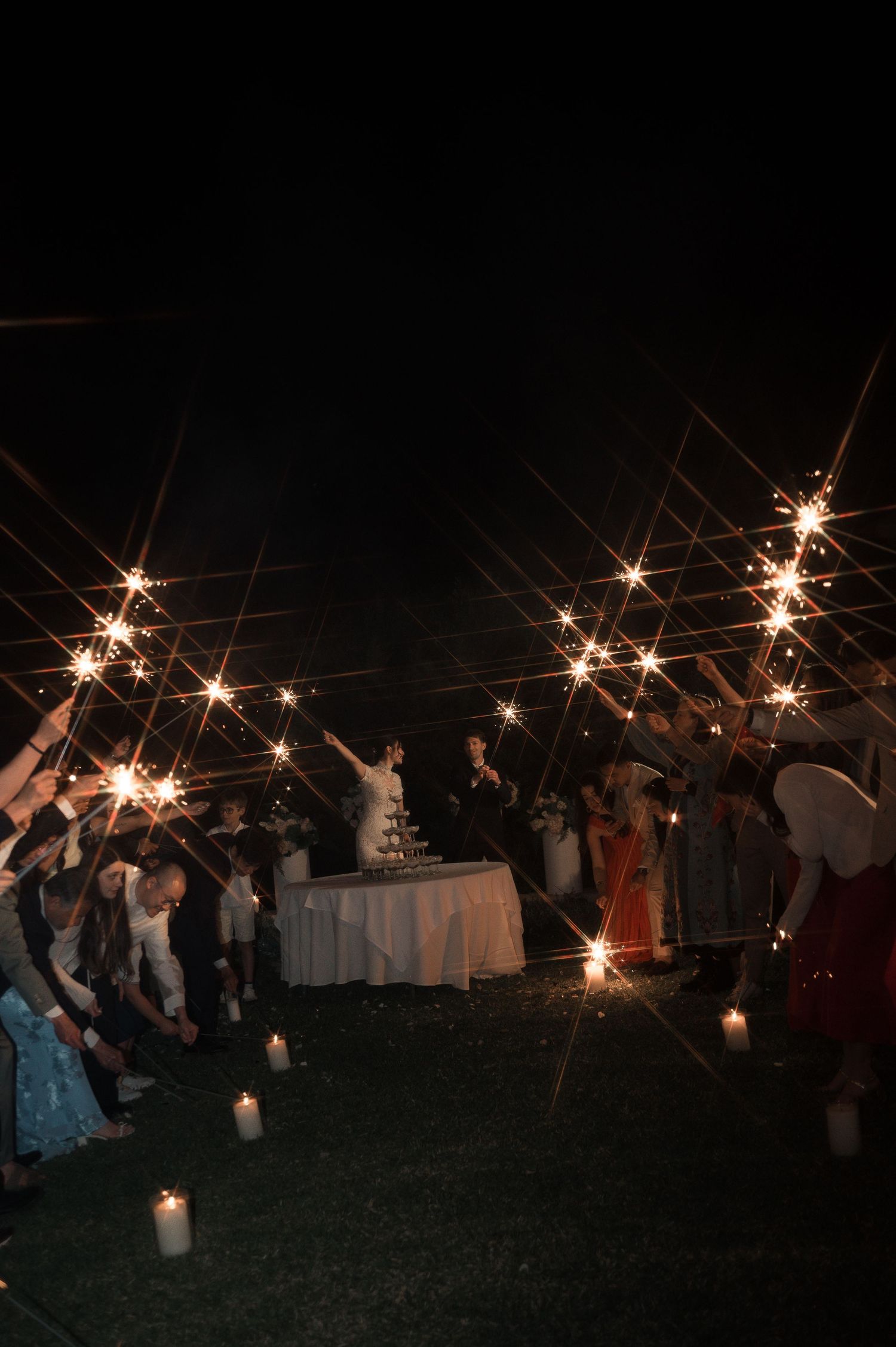 Guests holding sparklers in a circle around a wedding cake table at an outdoor nighttime celebration.
