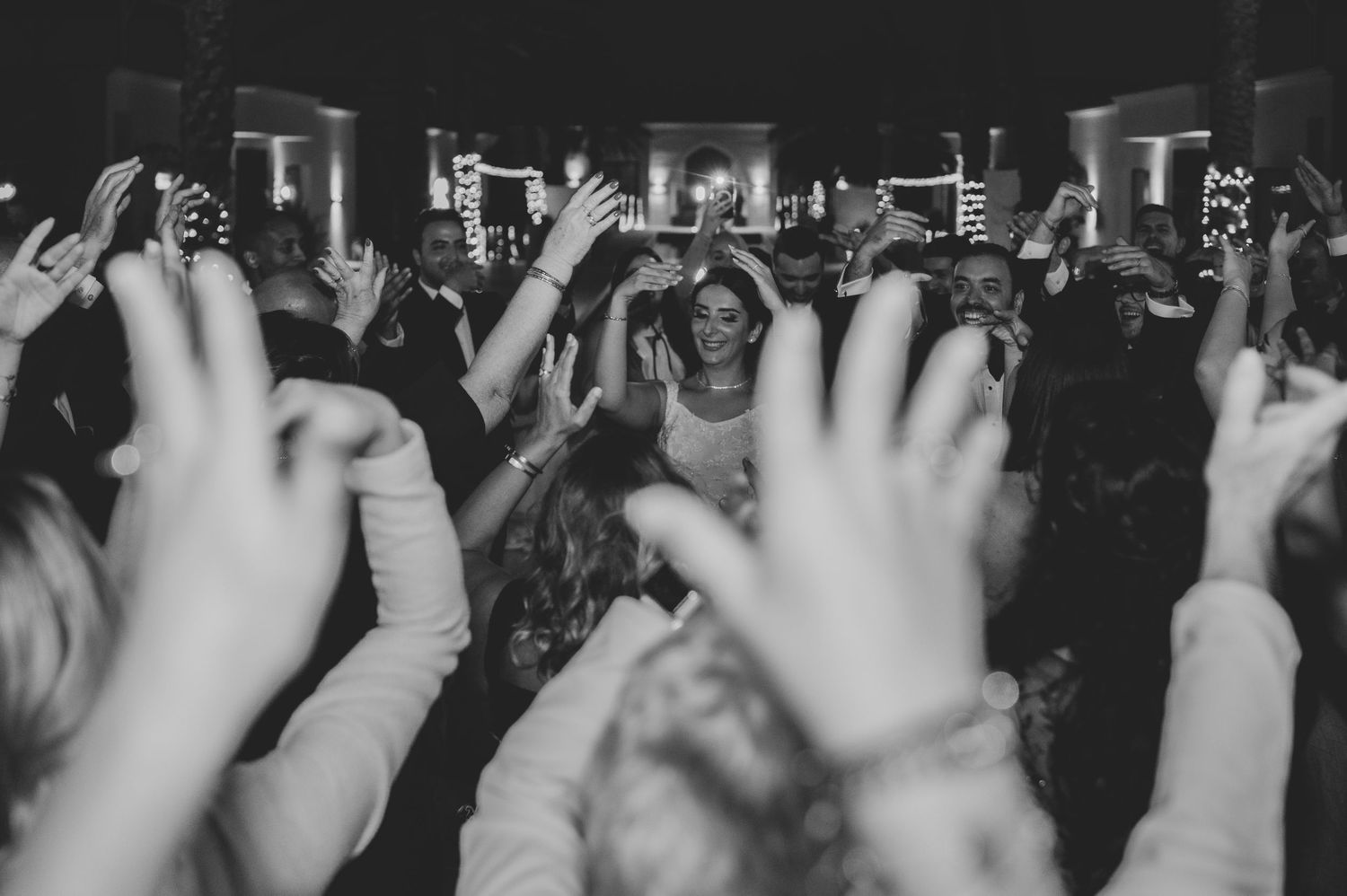 Black and white photo of bride surrounded by cheering guests with raised hands at wedding reception.