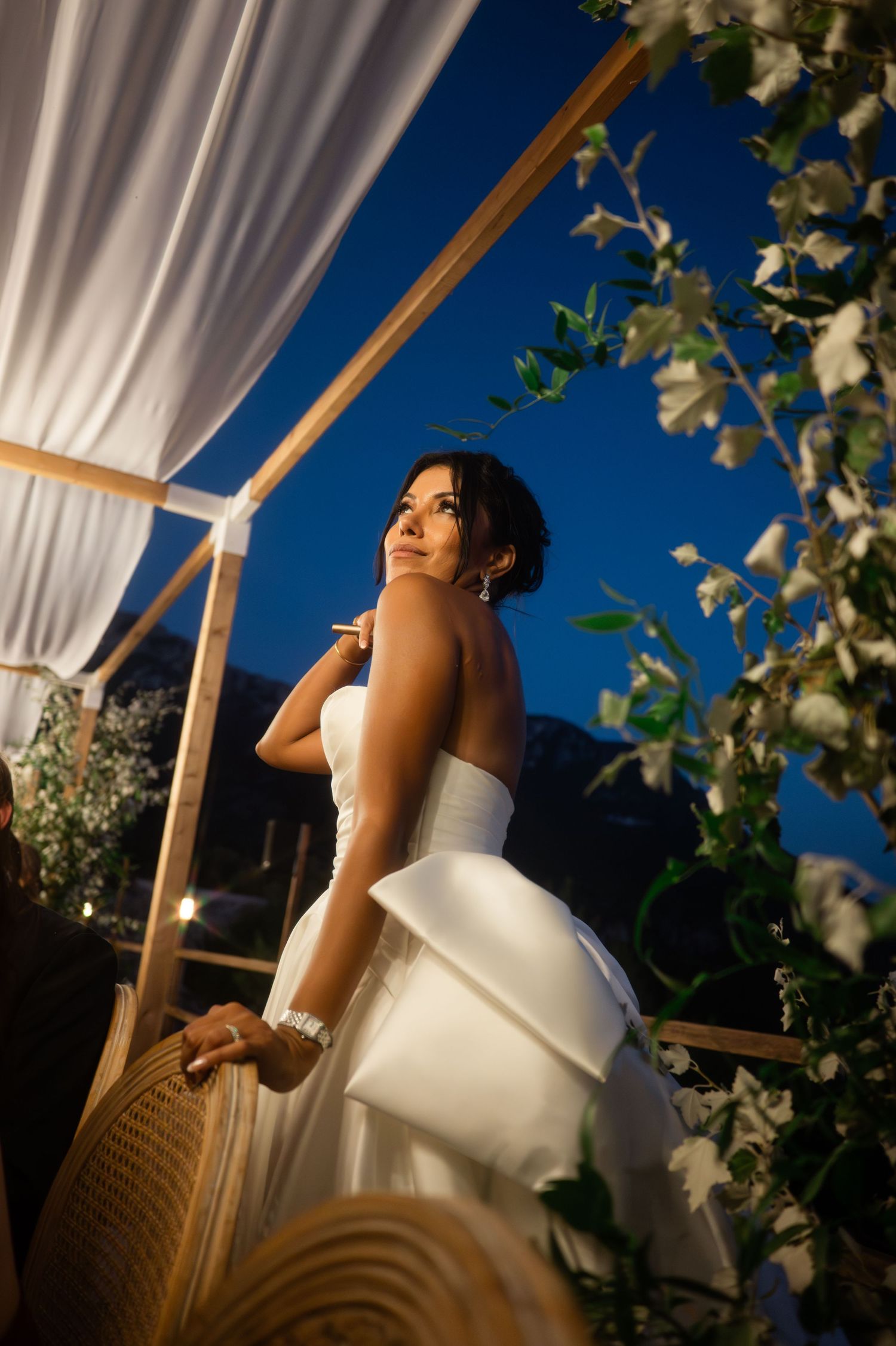 Beautiful bride in strapless white gown gazing upward under floral arch against deep blue night sky.