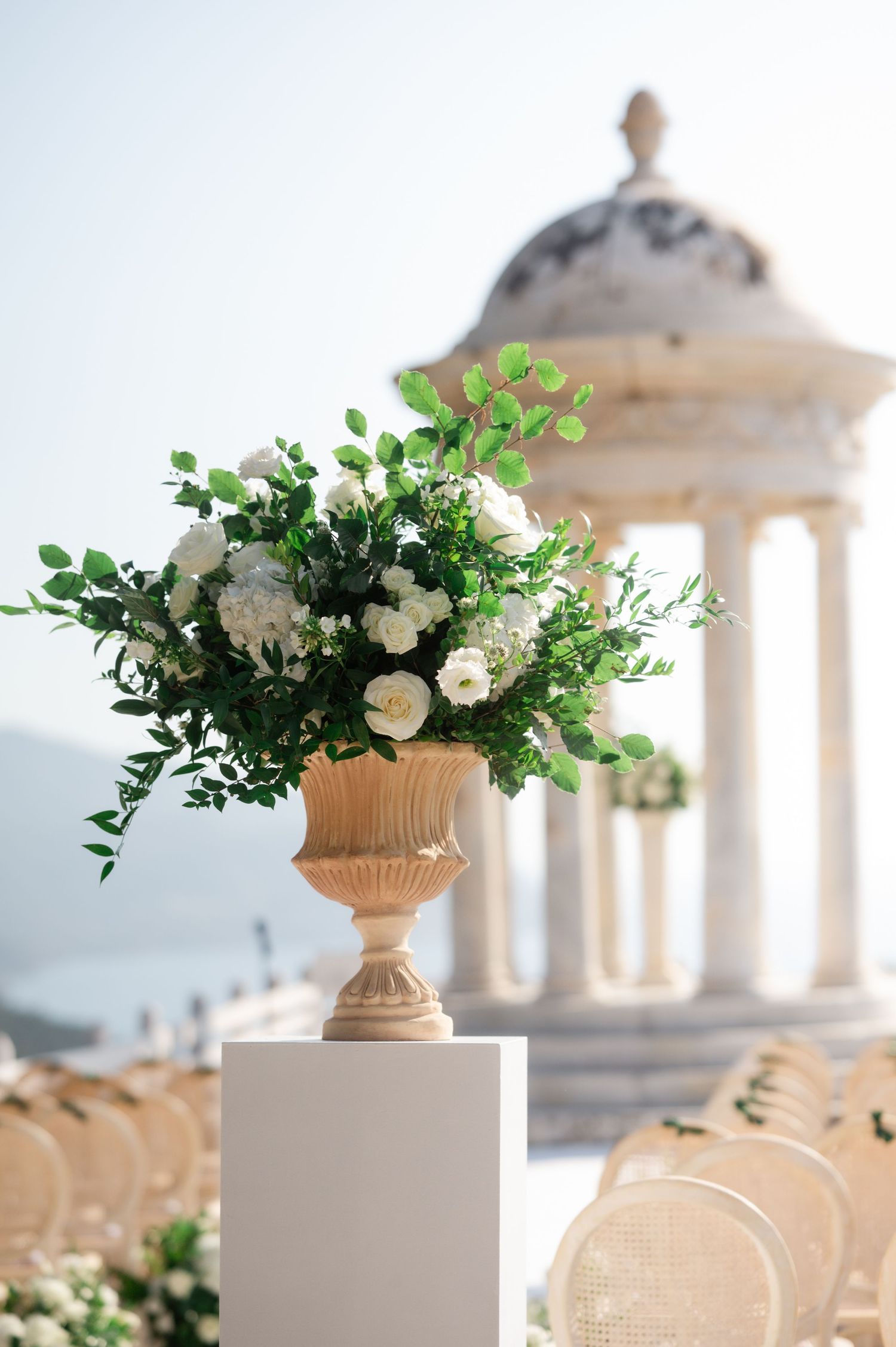 Elegant white floral arrangement in stone urn with classical rotunda backdrop at outdoor ceremony.