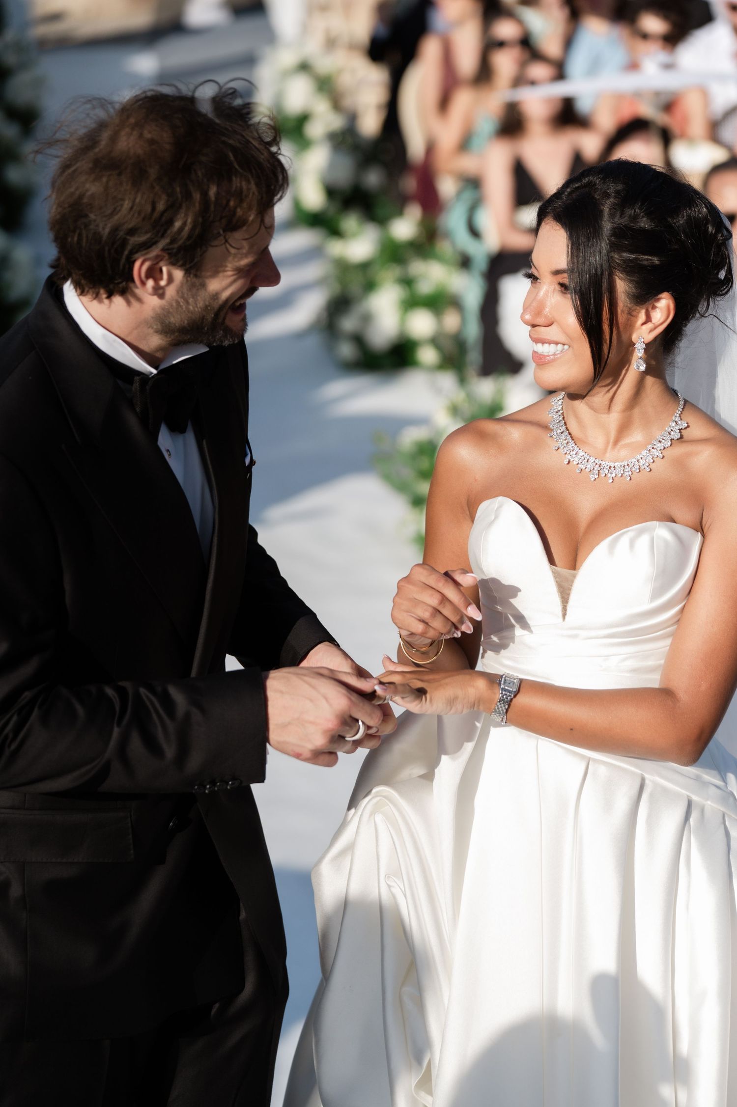 Joyful bride smiles as groom places wedding ring on her finger during outdoor ceremony.
