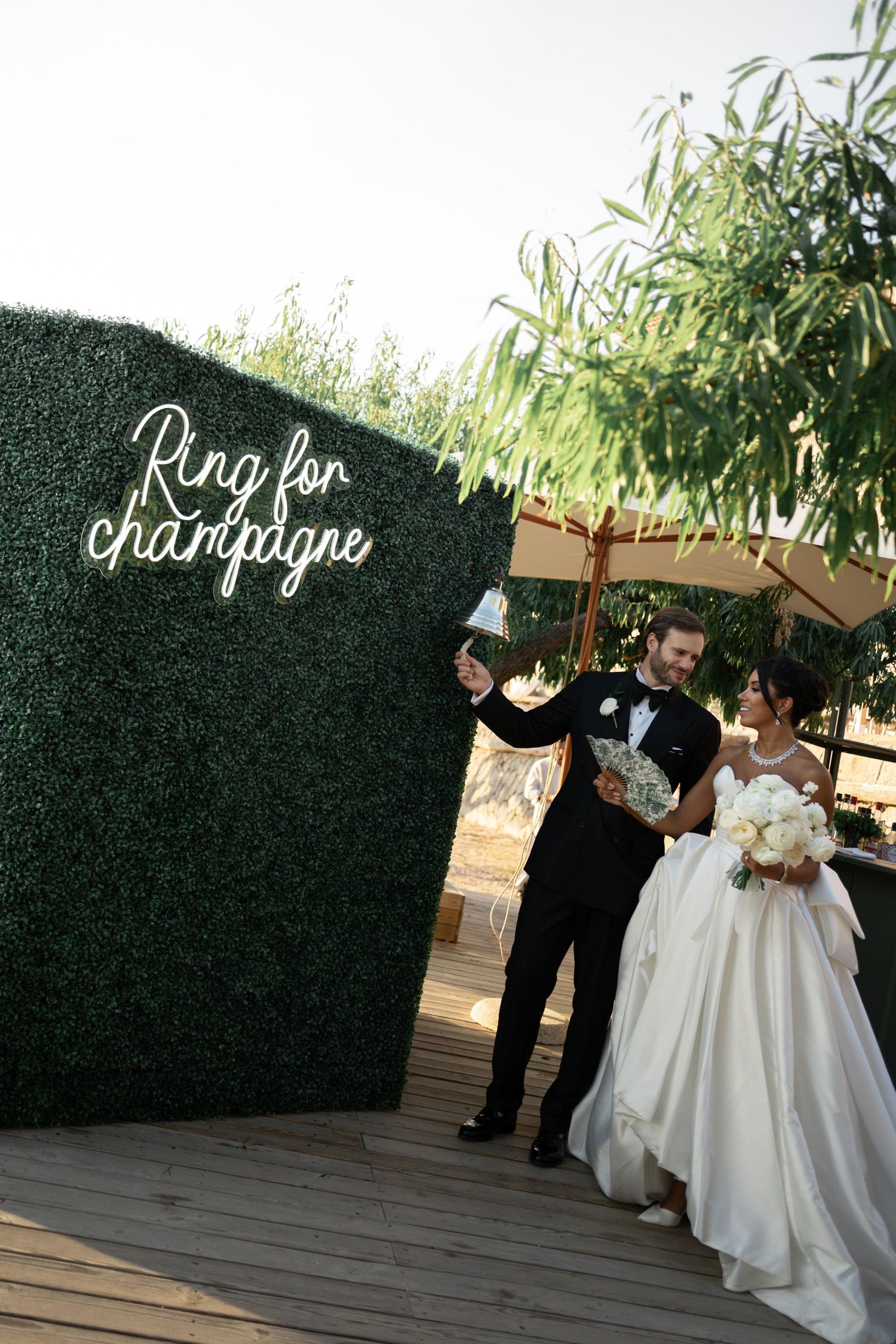Bride and groom ringing a bell at a green hedge wall with Ring for Champagne neon sign at their wedding.