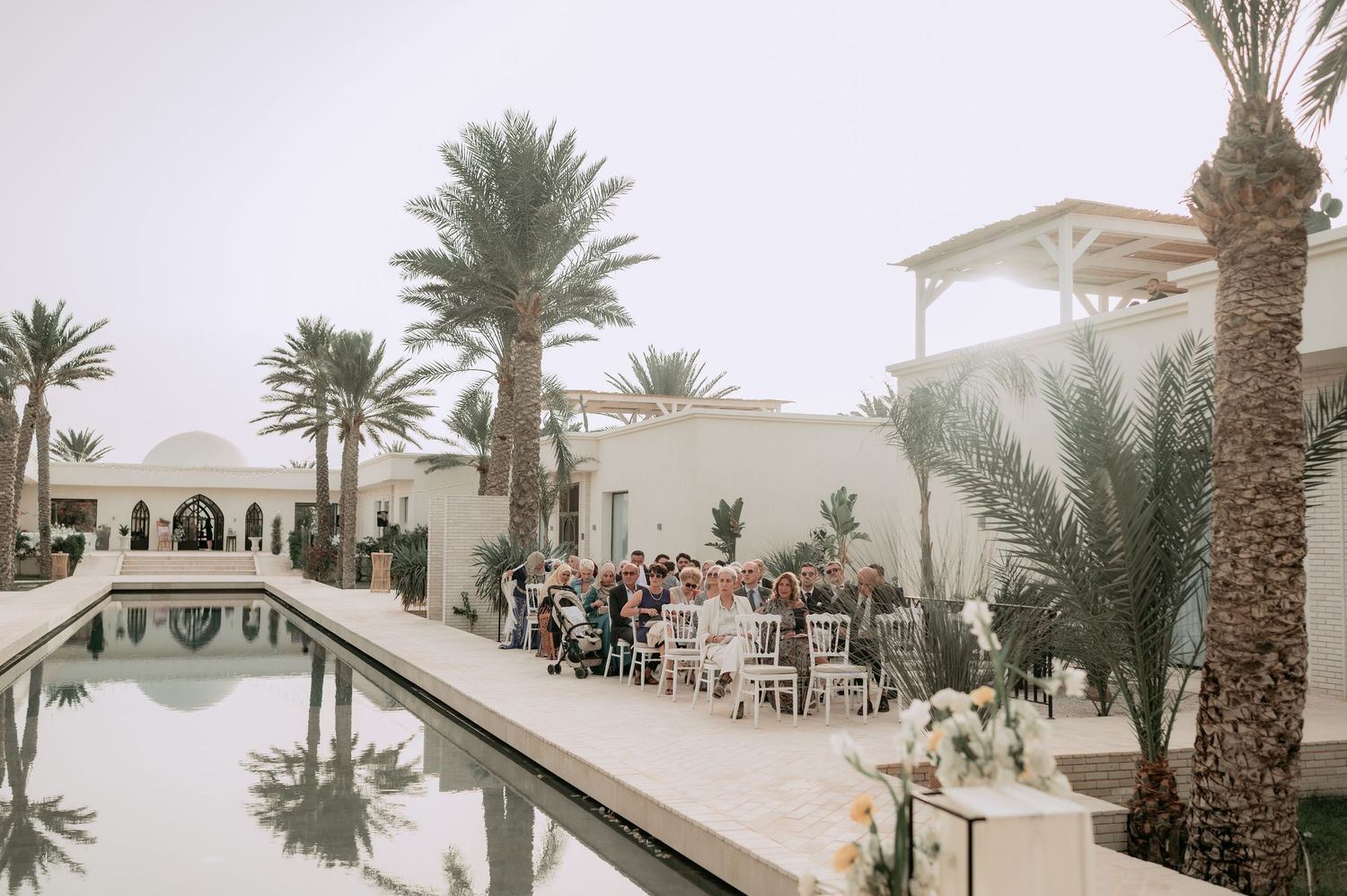 Outdoor wedding ceremony beside reflecting pool surrounded by palm trees at elegant white venue.