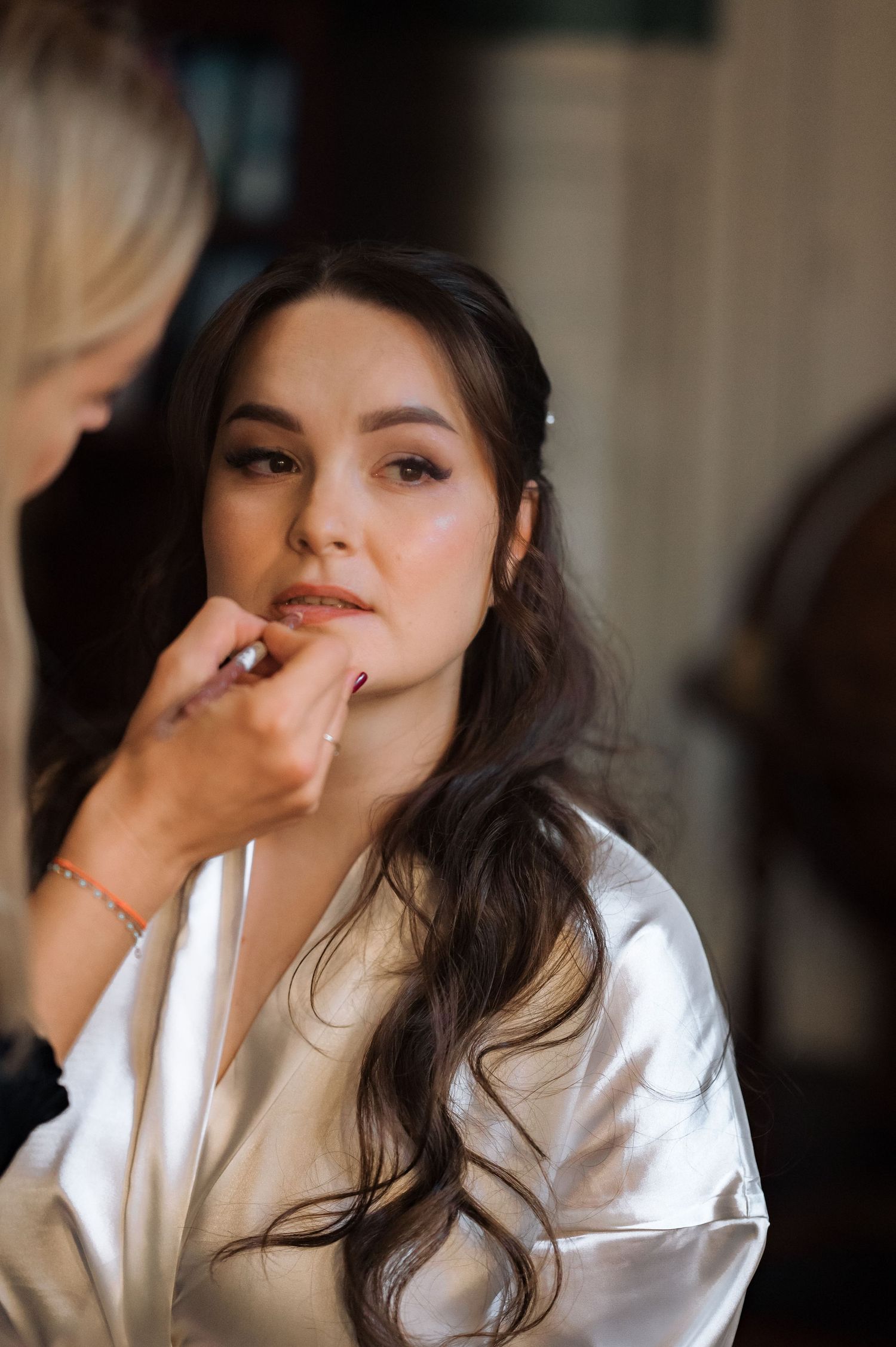 Makeup artist applying finishing touches to a brunette bride in a white satin robe on her wedding day.