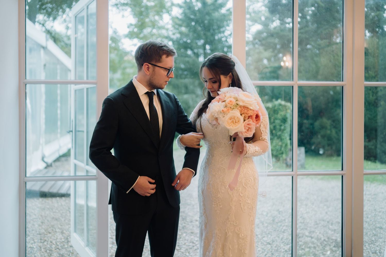 Groom adjusts bride's lace wedding dress sleeve near large glass doors in soft light.