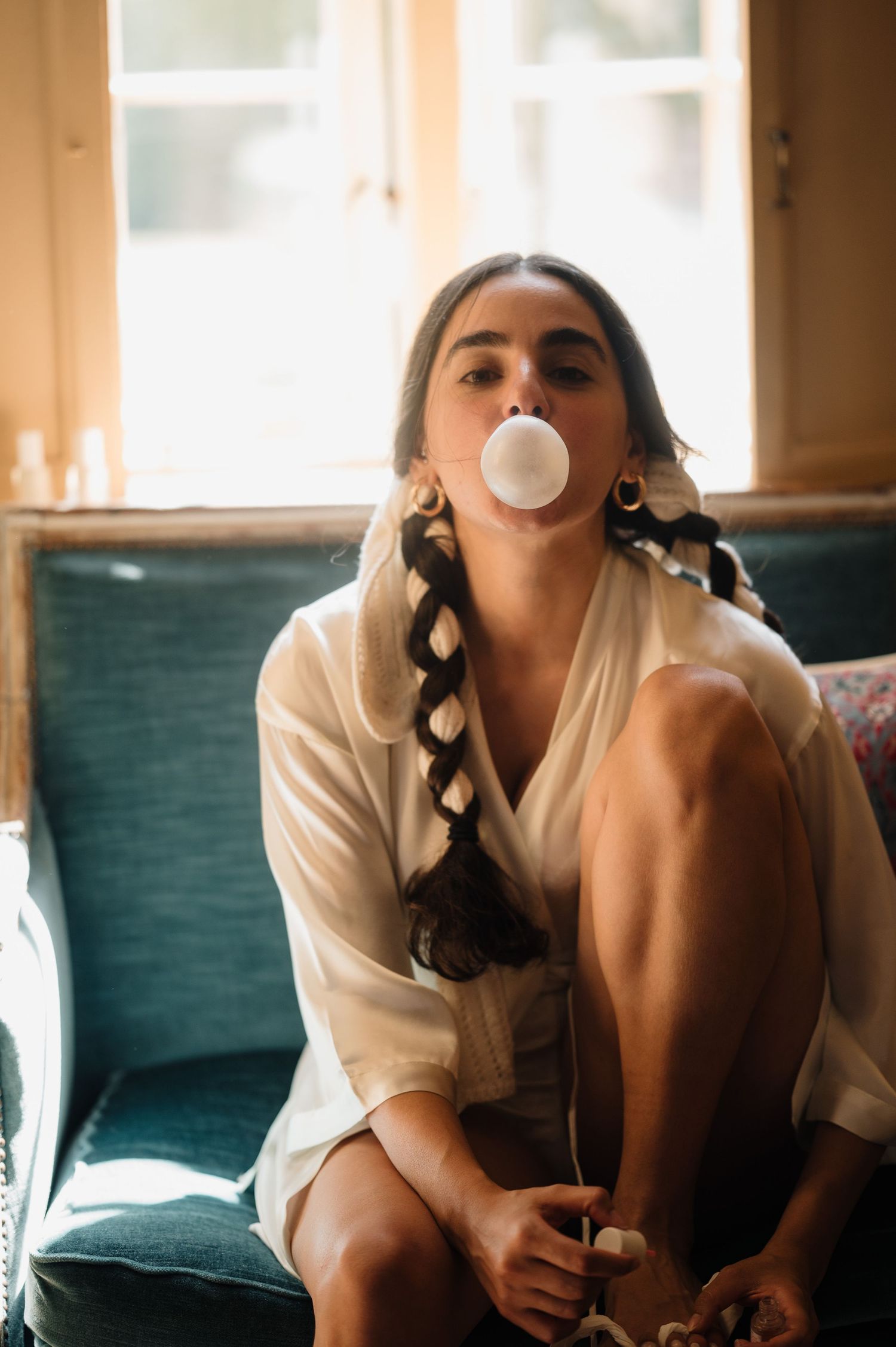 Young woman with braided hair blowing bubble gum bubble while sitting on teal sofa.