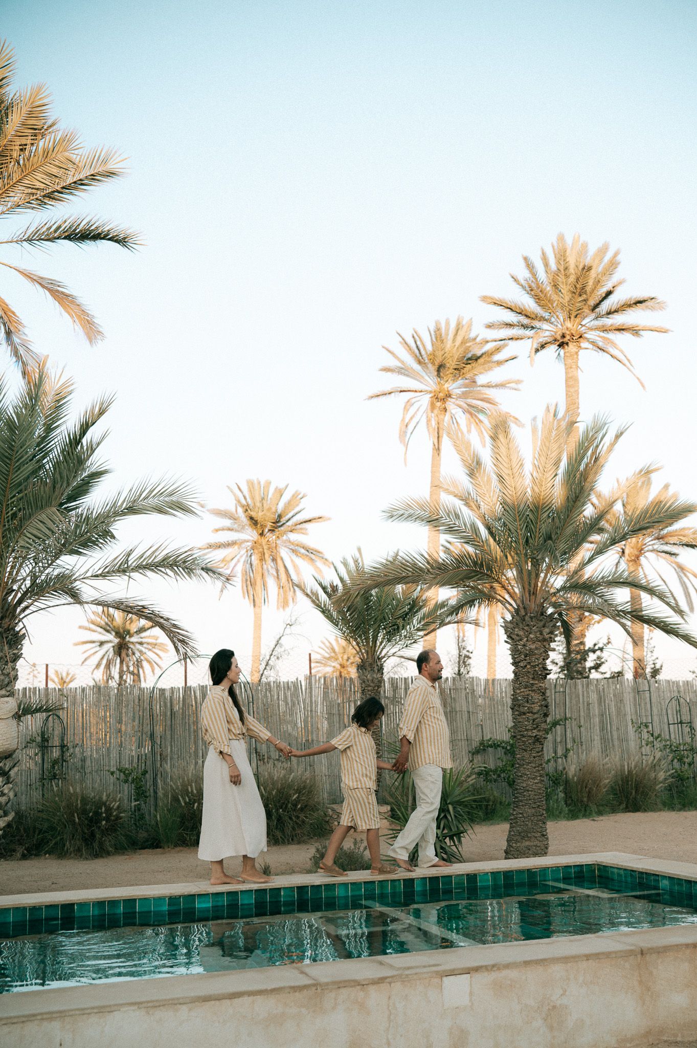 Family of three walking hand in hand beside pool surrounded by tall palm trees at sunset.