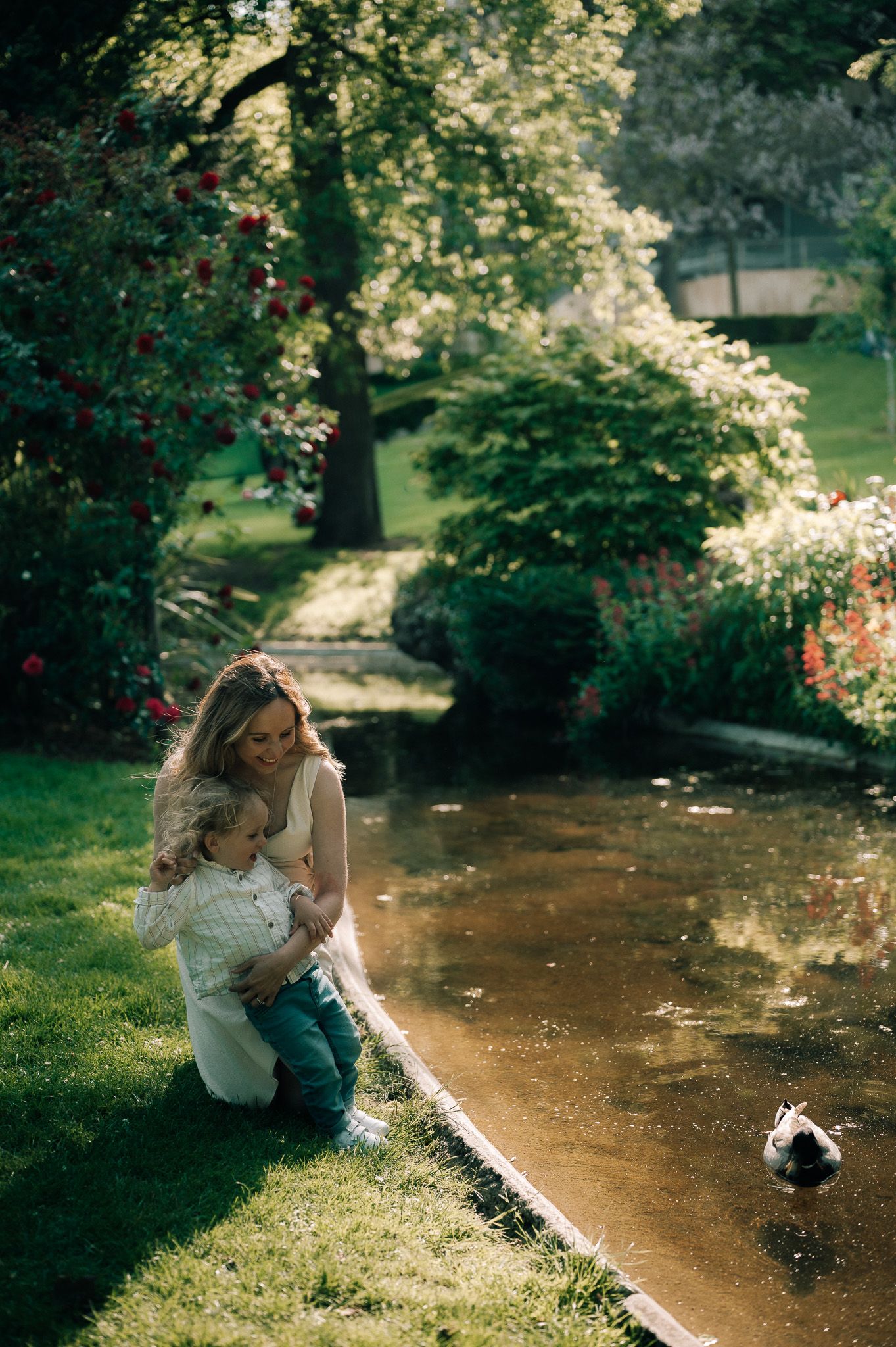 Mother kneeling by a sunlit garden pond, tenderly holding her toddler while watching a duck swim by.