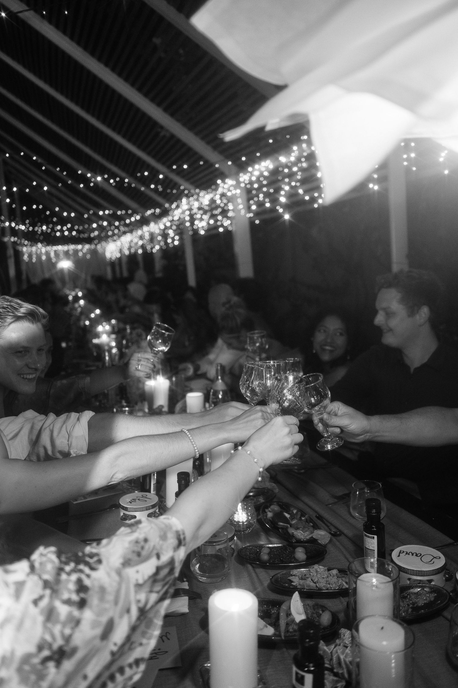 Wedding guests clink glasses in a toast under warm string lights at night reception.