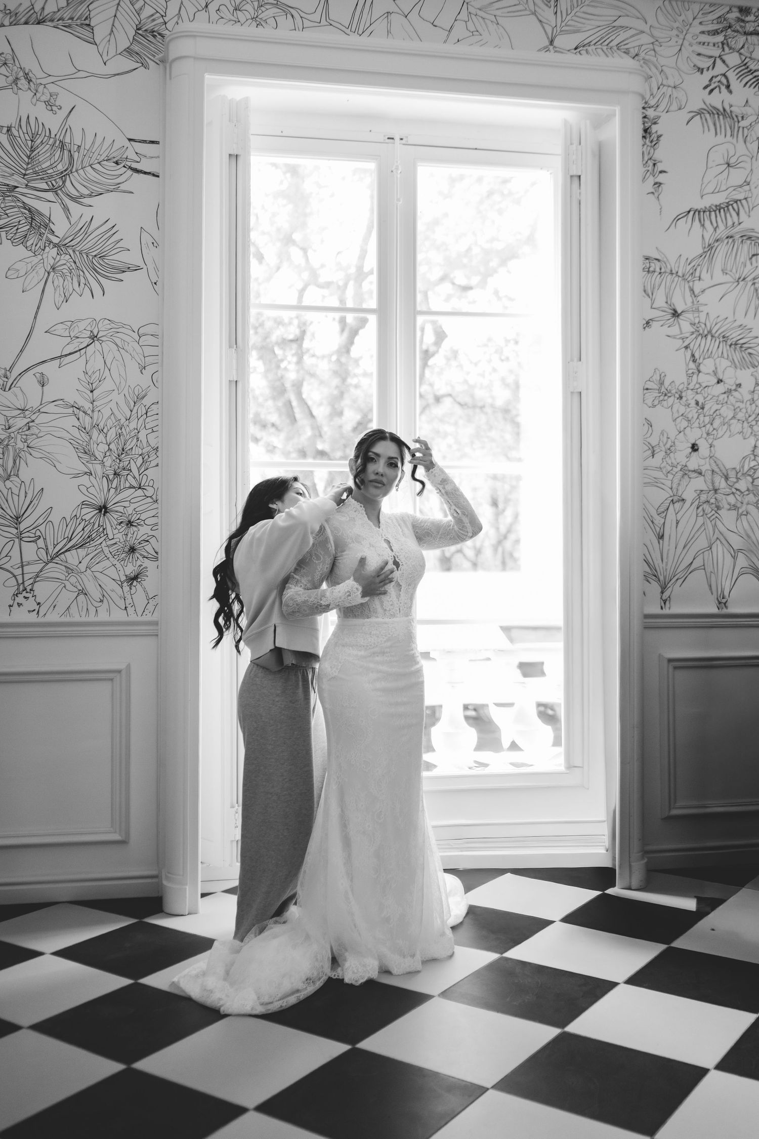 Black and white photo of bridesmaid helping bride get ready by bright window.