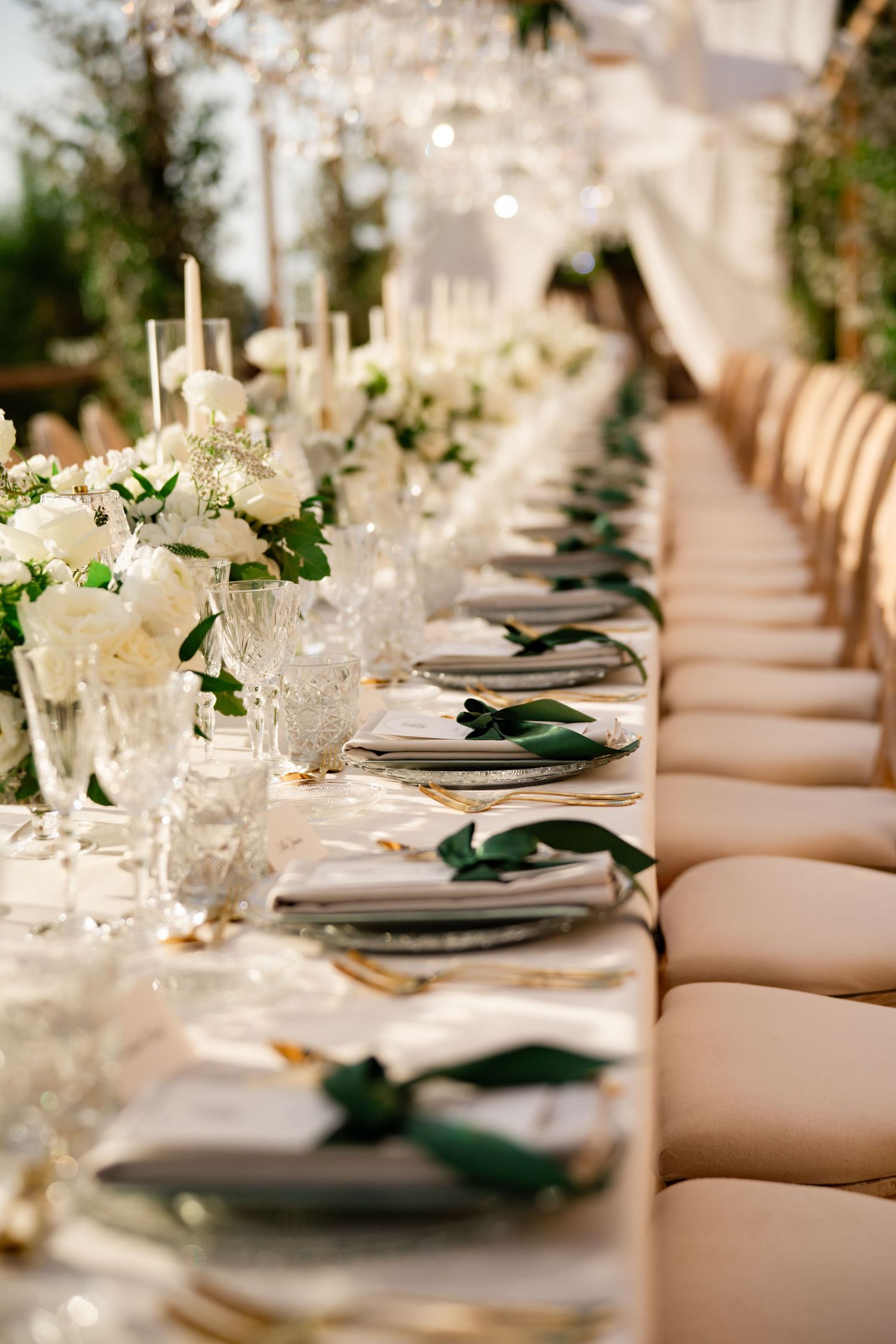 Long wedding reception table with white roses, crystal glassware, gold cutlery, green ribbon napkins, and tall candles.