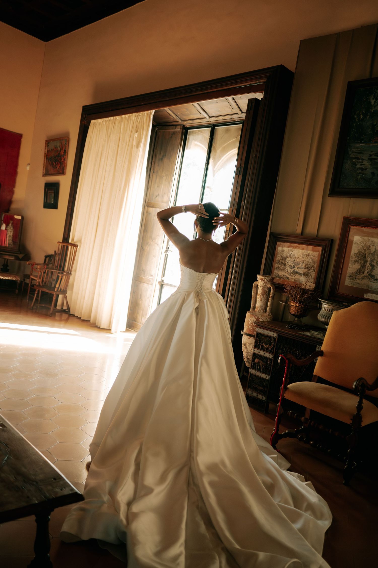 Bride in backless white ball gown with long train stands before ornate mirror in vintage room.