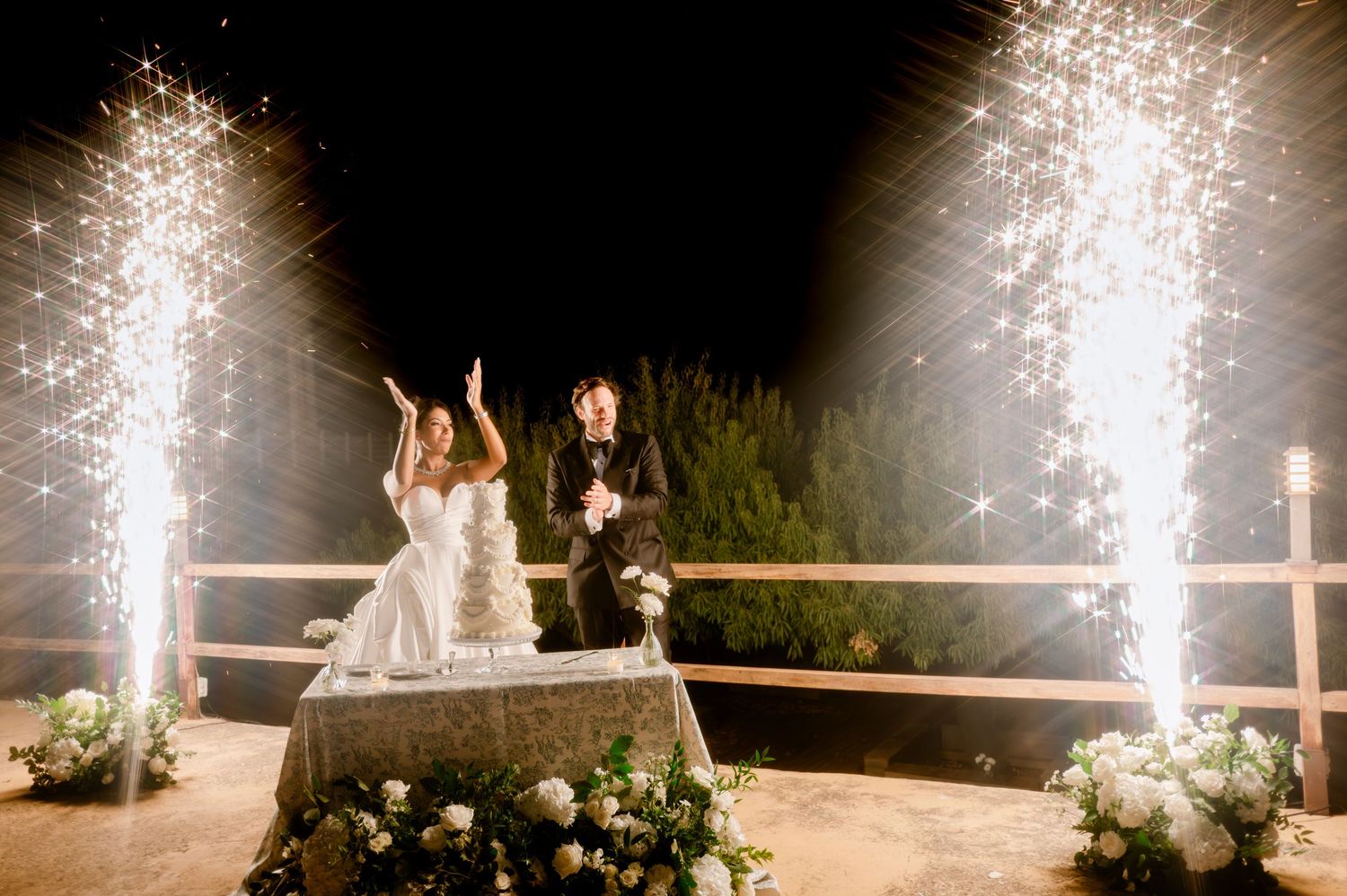 Newlyweds stand before stunning twin fountain sparklers during outdoor wedding cake celebration at night.