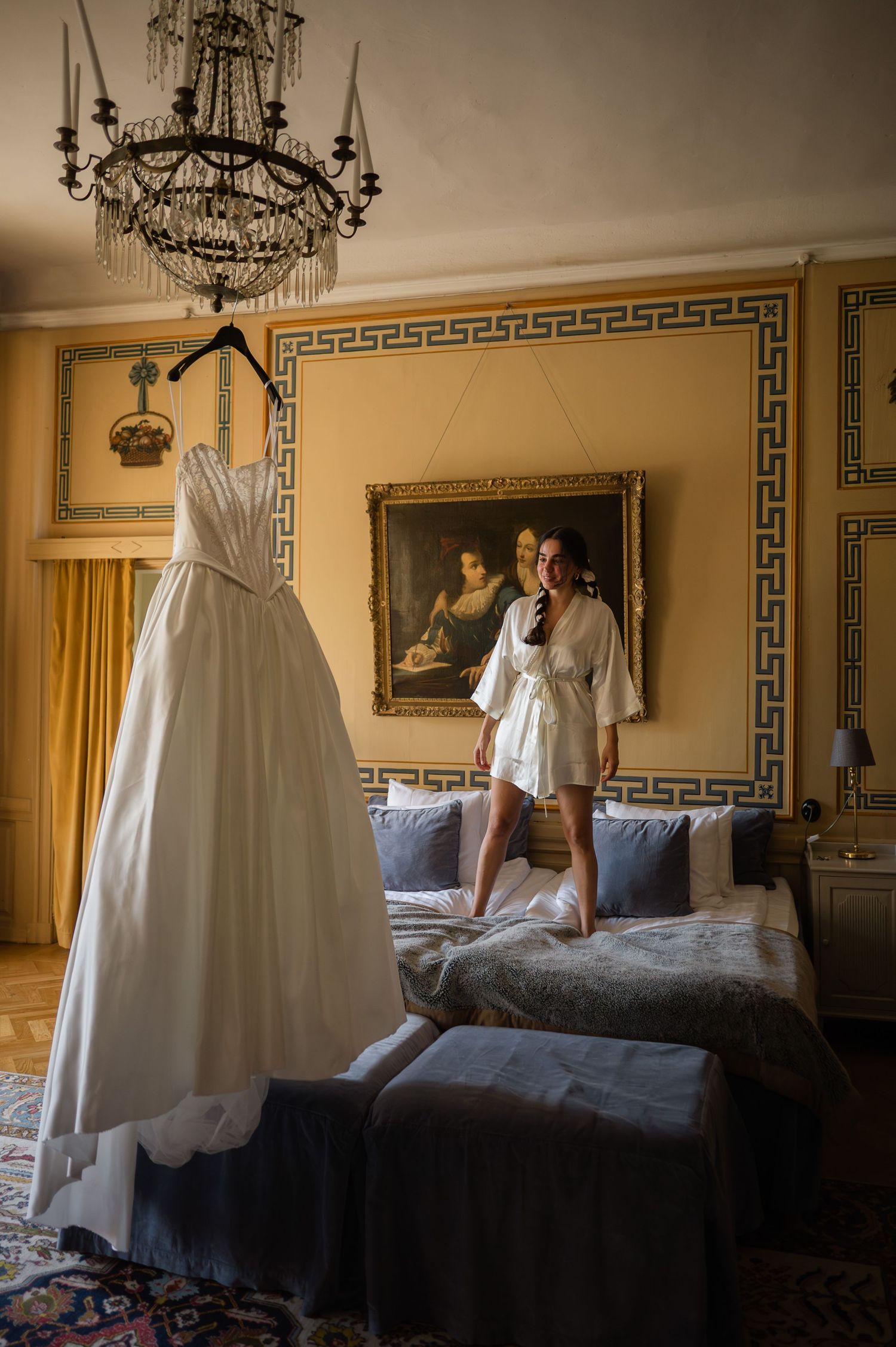 Bride in white robe standing on bed admiring wedding gown hanging in ornate gold-accented hotel room.