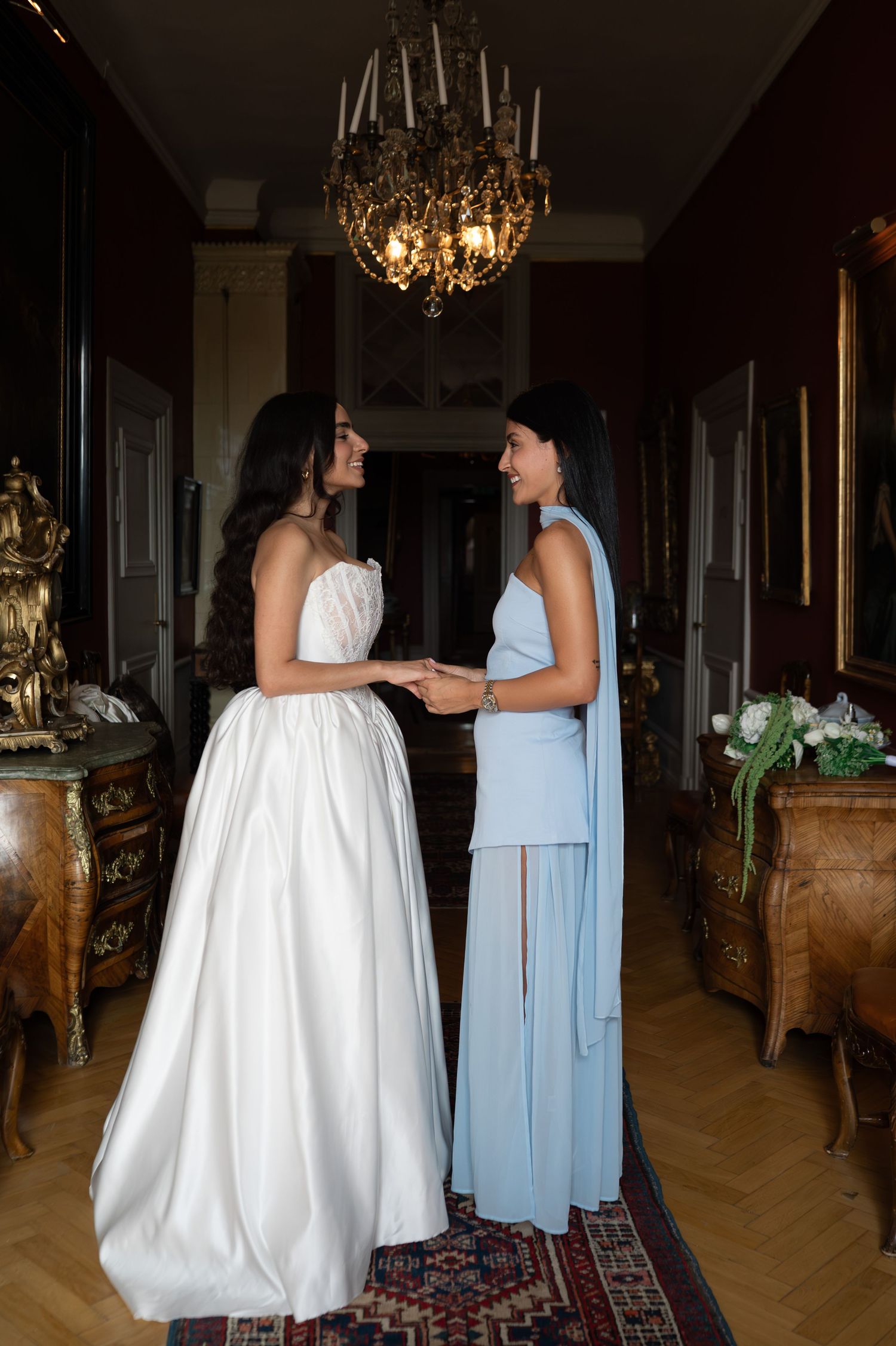 Bride and bridesmaid in blue dress holding hands and smiling in elegant chandelier-lit hallway.