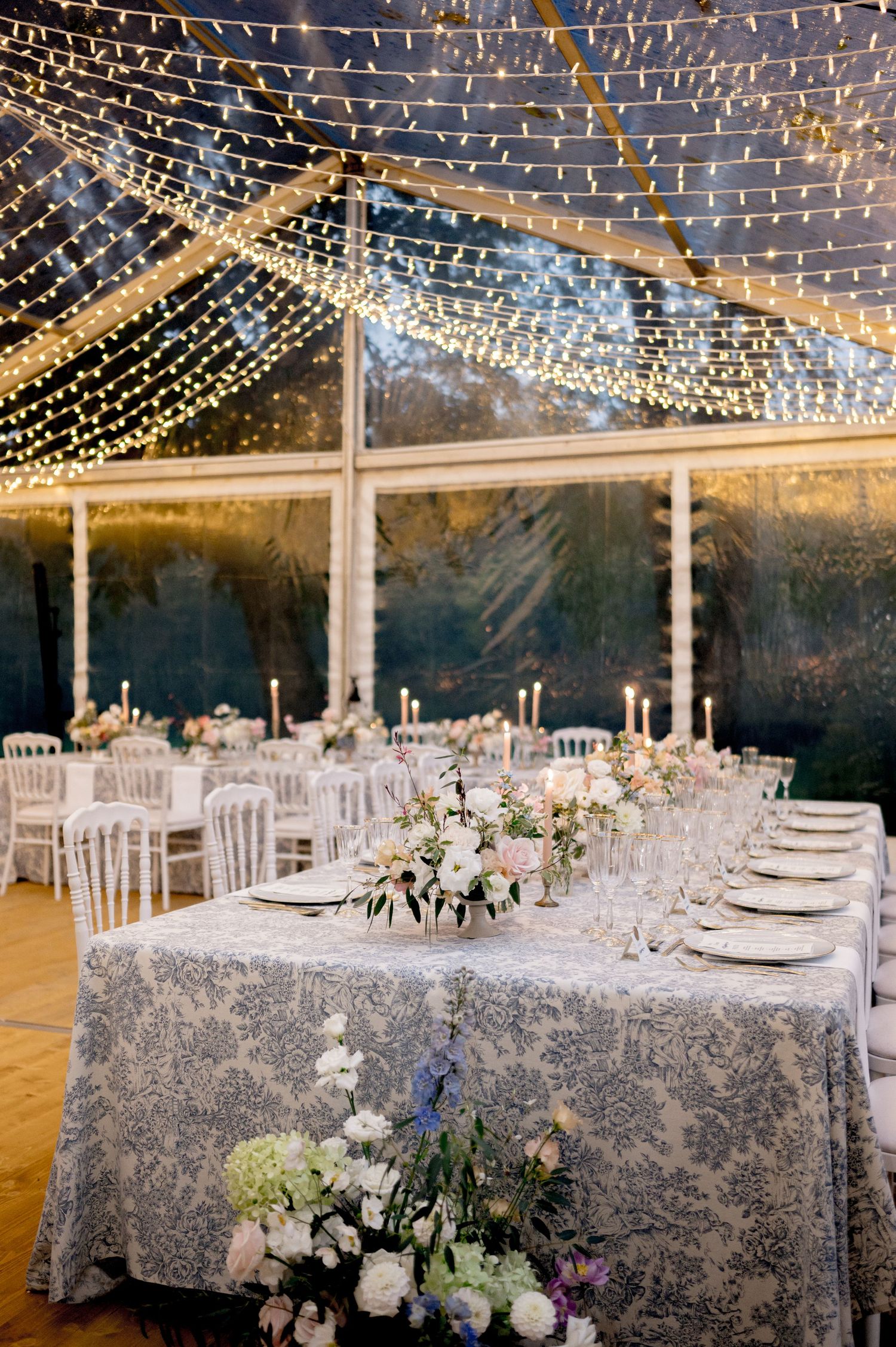 Romantic wedding reception under fairy light canopy with blue toile tables, white chairs, and floral centerpieces.