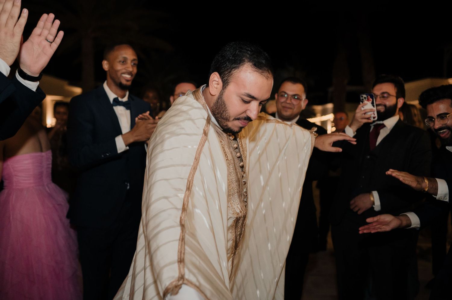 Groom in white traditional Moroccan djellaba celebrated by guests at wedding reception.