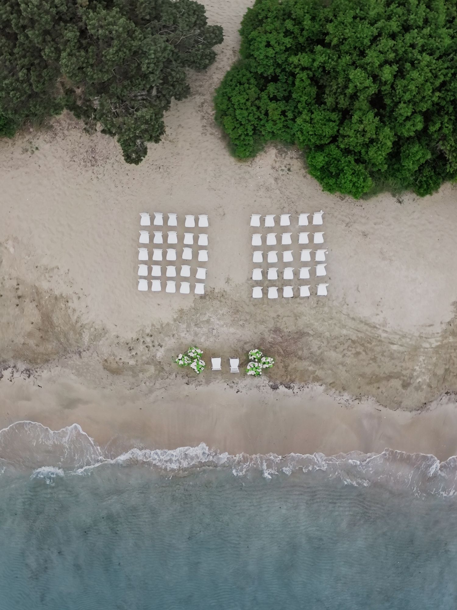 Aerial view of white wedding ceremony chairs arranged in rows on a sandy beach beside turquoise water.