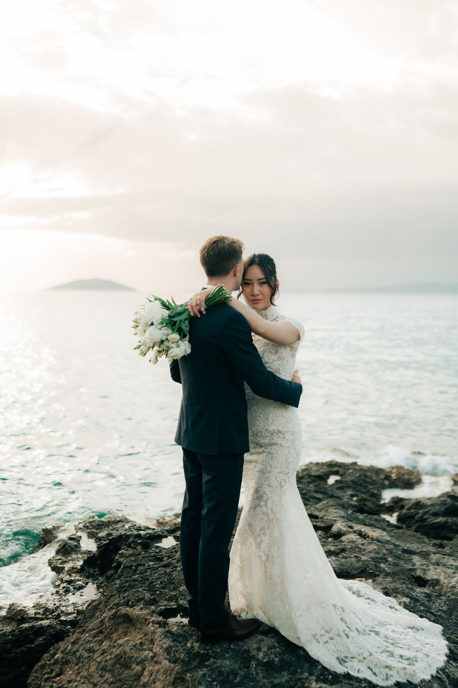 Bride and groom embracing on rocky seaside cliffs at sunset, bride holding white bouquet in lace mermaid gown.