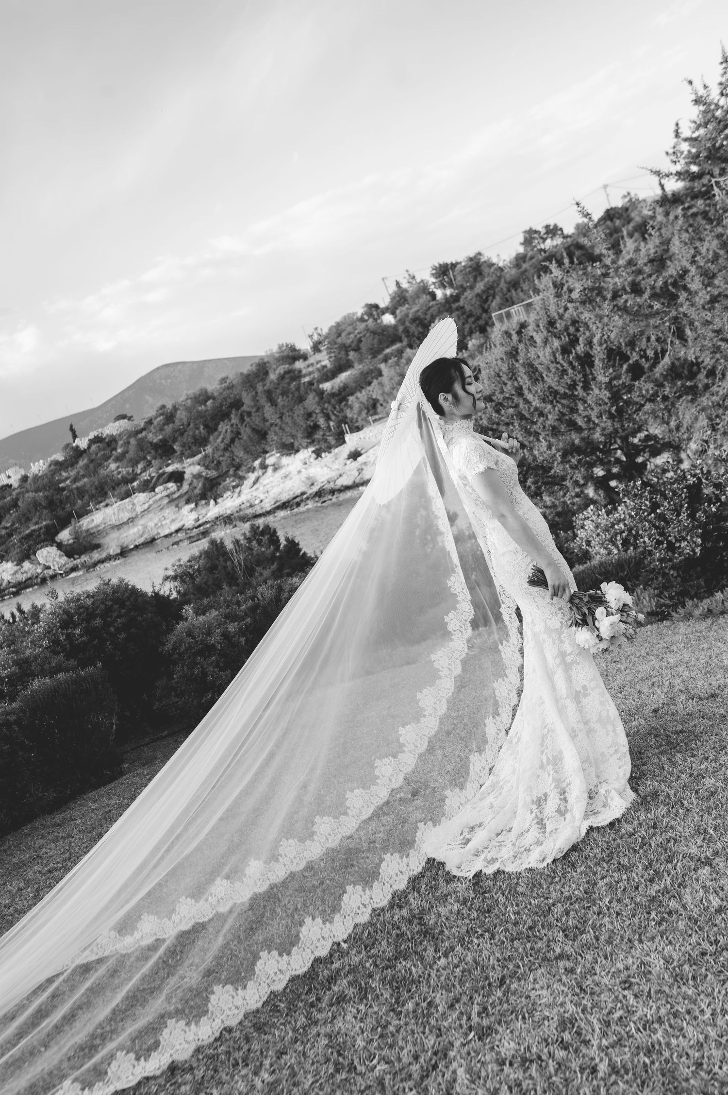Bride in lace gown with long cathedral veil trailing on green hillside landscape.