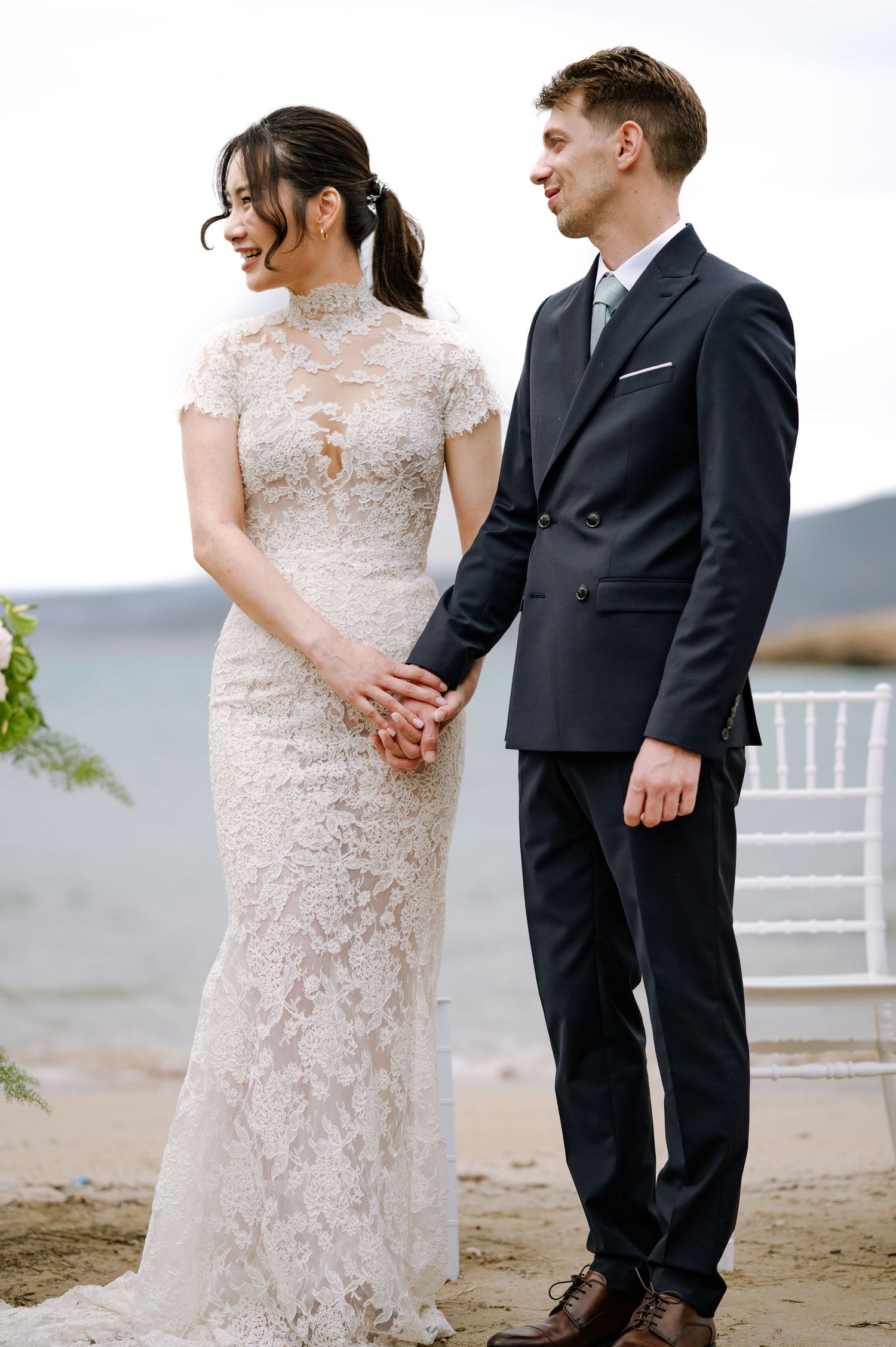 Bride in high-neck lace gown and groom in dark suit hold hands during beach wedding ceremony.