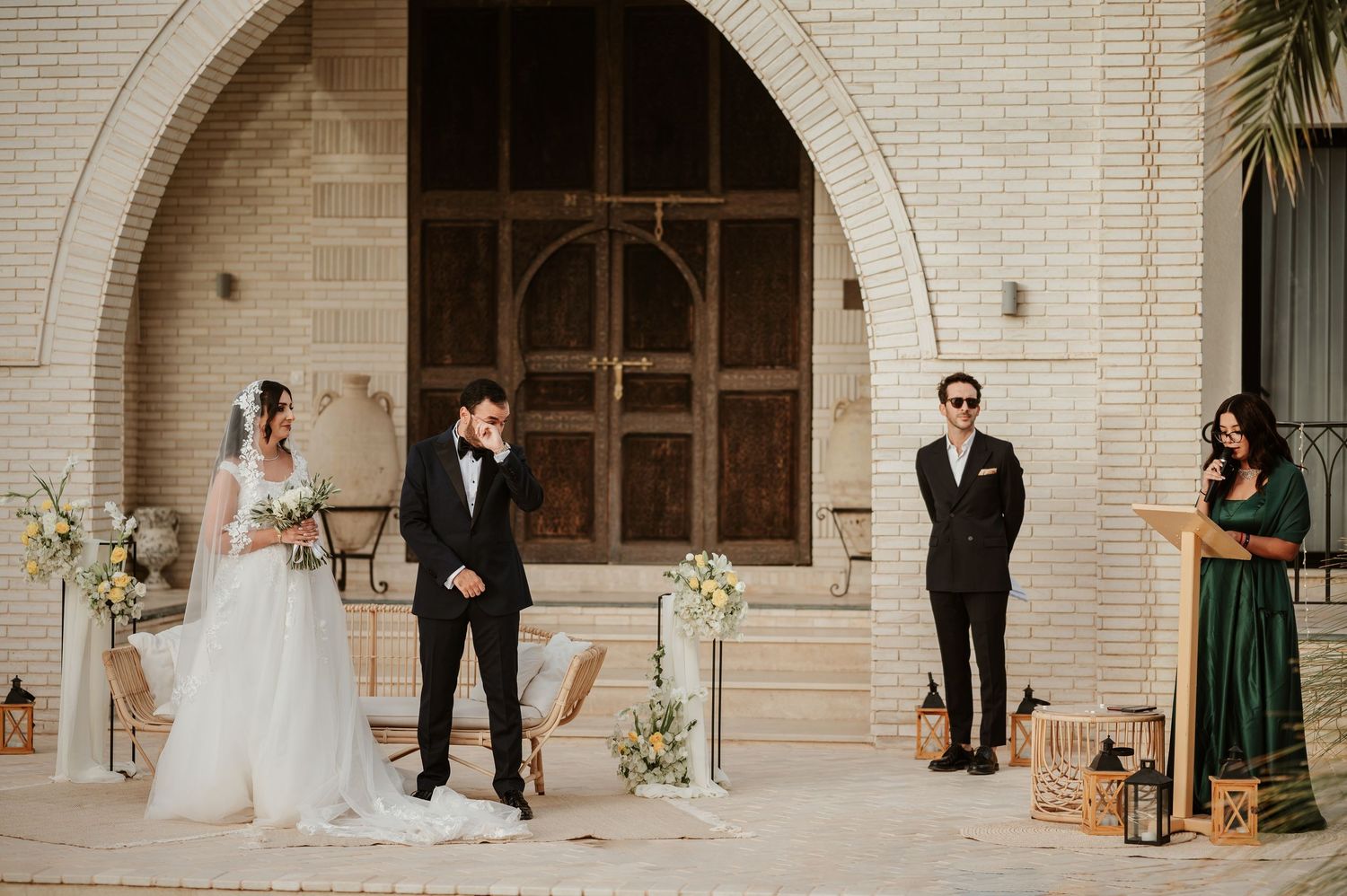 Bride and groom stand at altar before ornate wooden doors during outdoor ceremony with officiant in green.