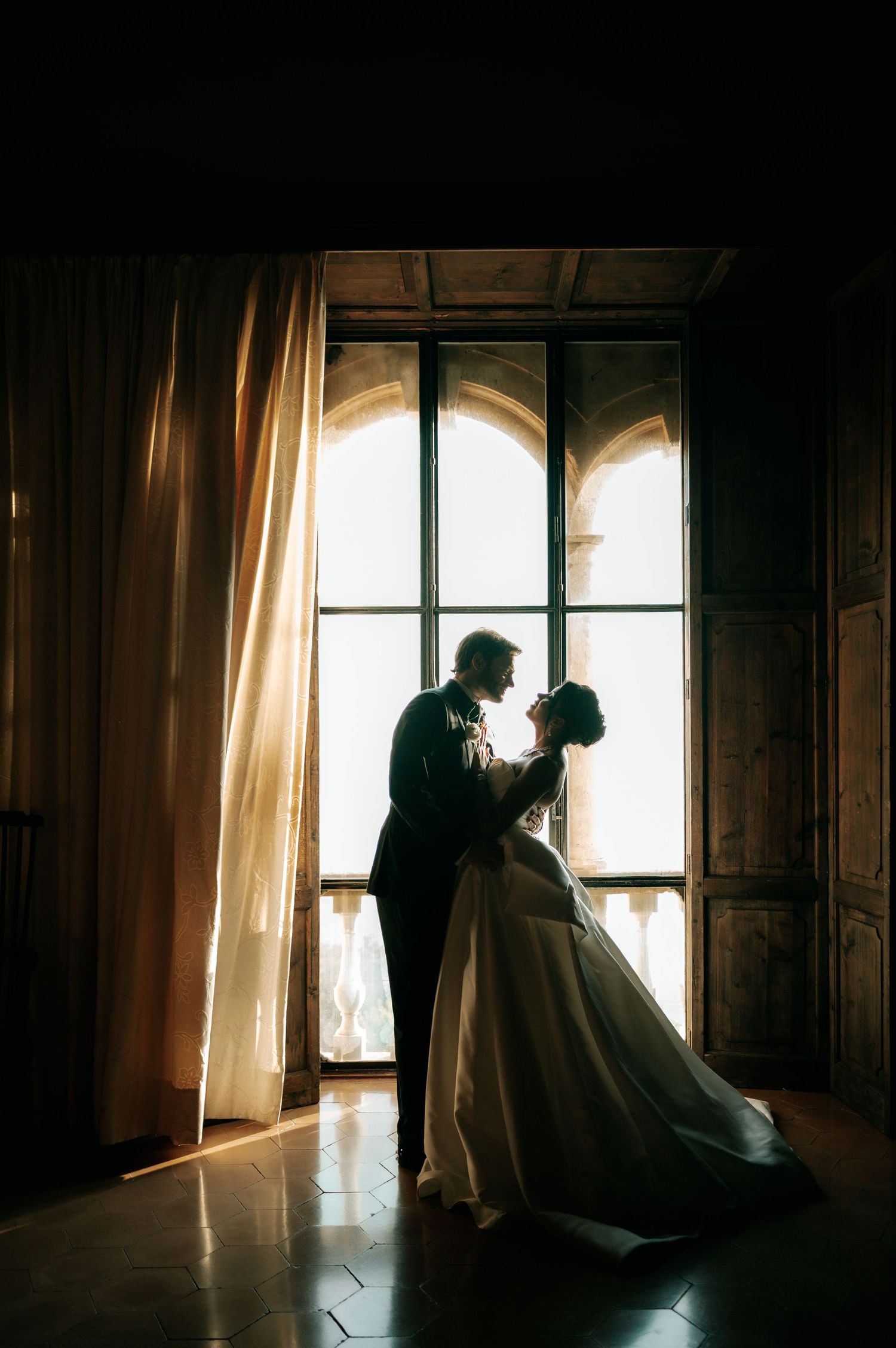 Bride and groom silhouetted against a glowing arched window in a dark elegant room.