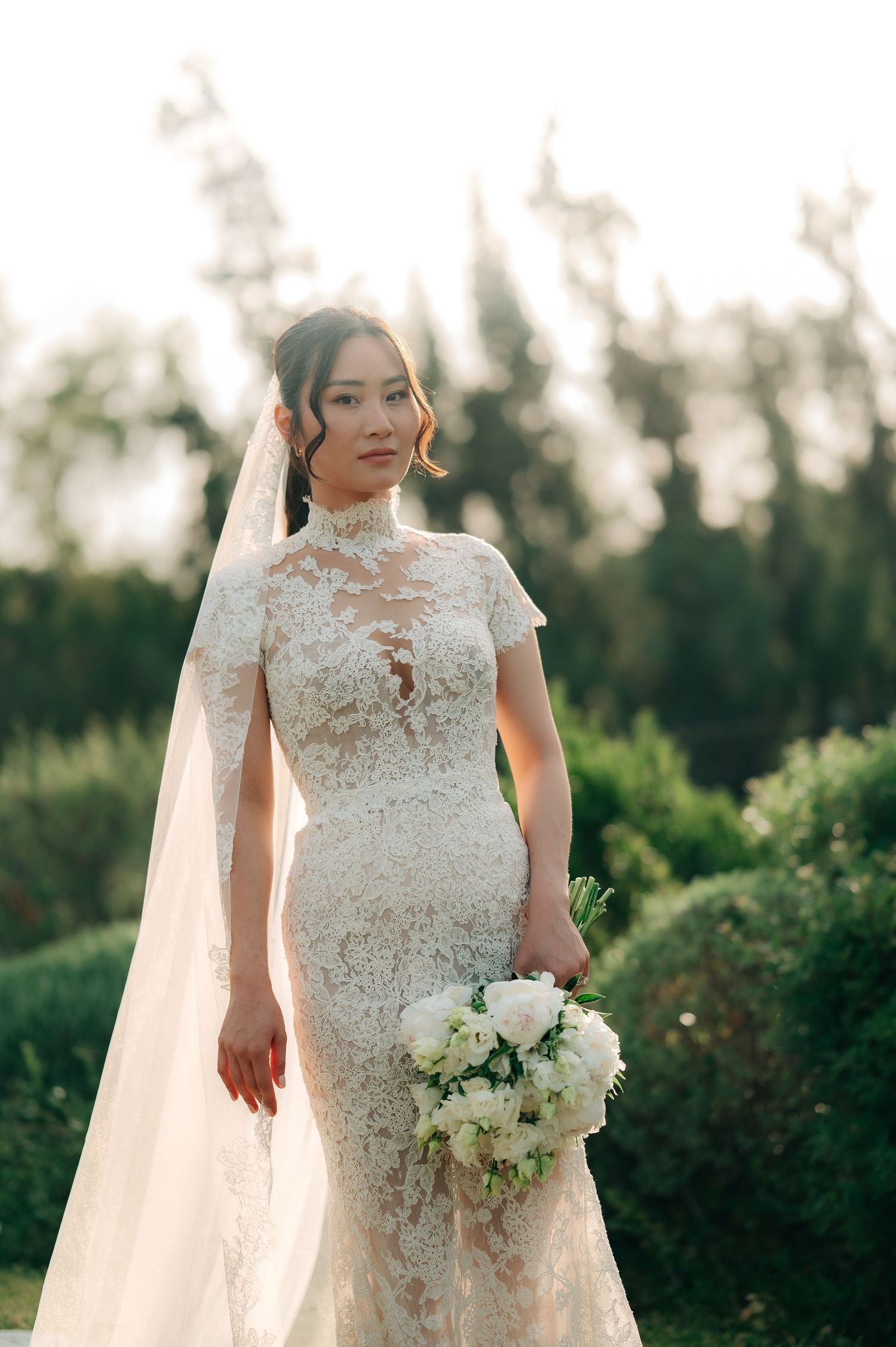 A bride in a lace high-neck gown with veil holds a white bouquet outdoors amid lush greenery.